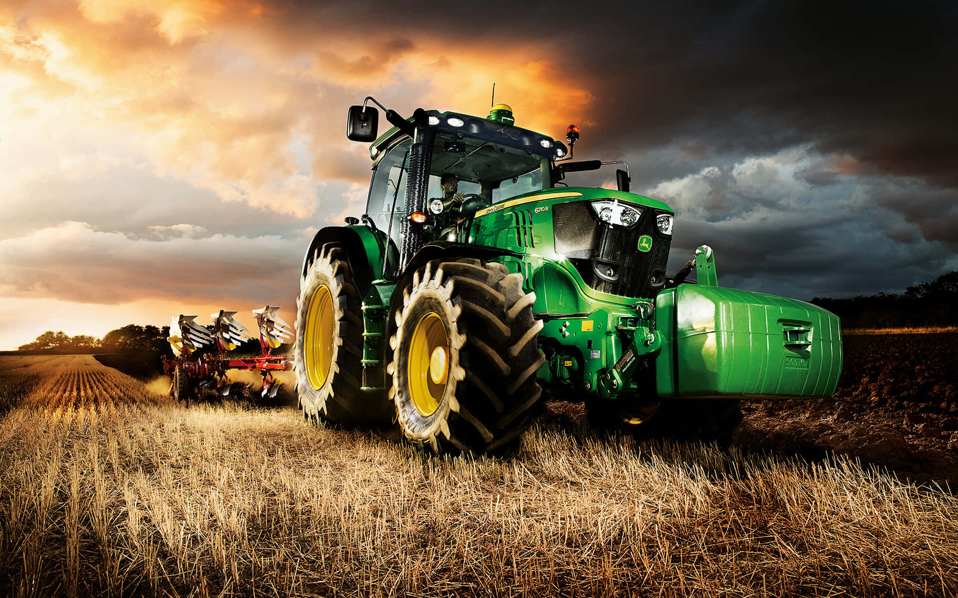 John Deere 桌布 壁紙 — 一輛綠色約翰迪爾拖拉機在烏雲密佈的戲劇性日落天空下犁著金色的田野。