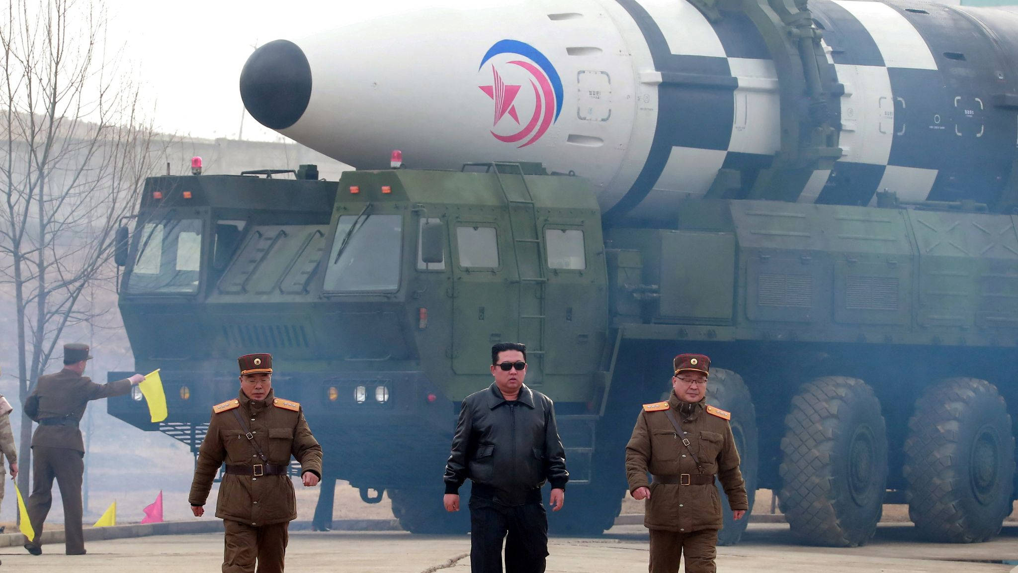 North Korea 桌布 壁紙 — 一輛綠色軍車上裝載著大型導彈，身穿制服的官員在陰天中走在車前。