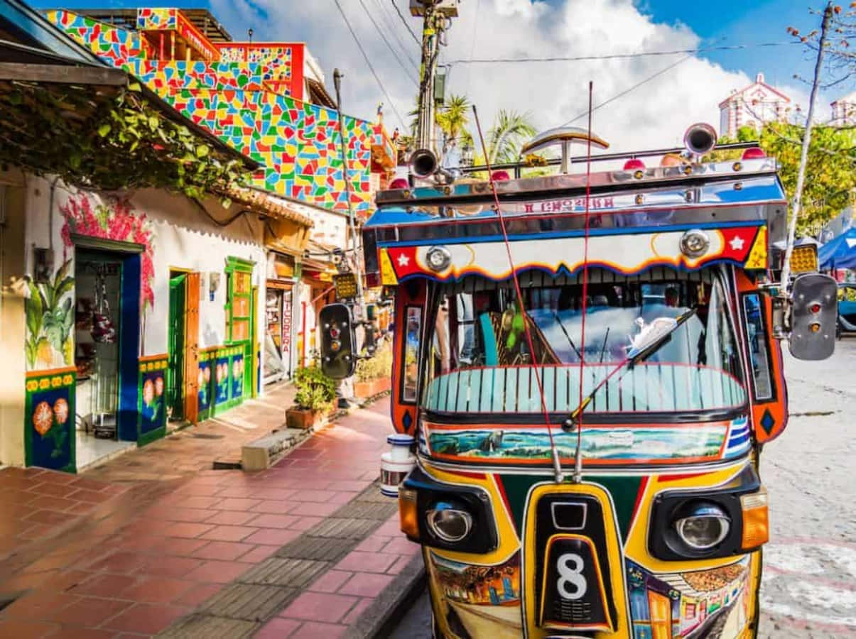 Colombia 桌布 壁紙 — 一輛色彩繽紛、帶有藝術塗鴉的自動人力車，在晴朗天空下，主宰著一條兩旁都是彩繪建築的熱鬧街道。