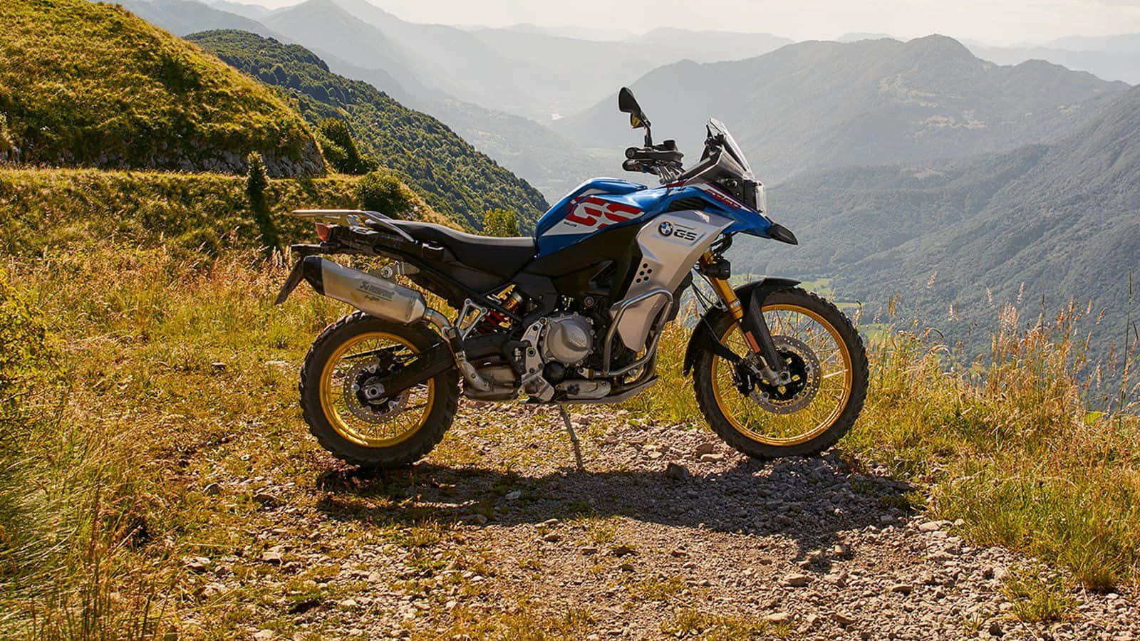 Thrill 桌布 壁紙 — 一輛藍白色的BMW GS冒險電單車停泊在岩石山徑上，俯瞰著陽光下起伏的綠色山丘。