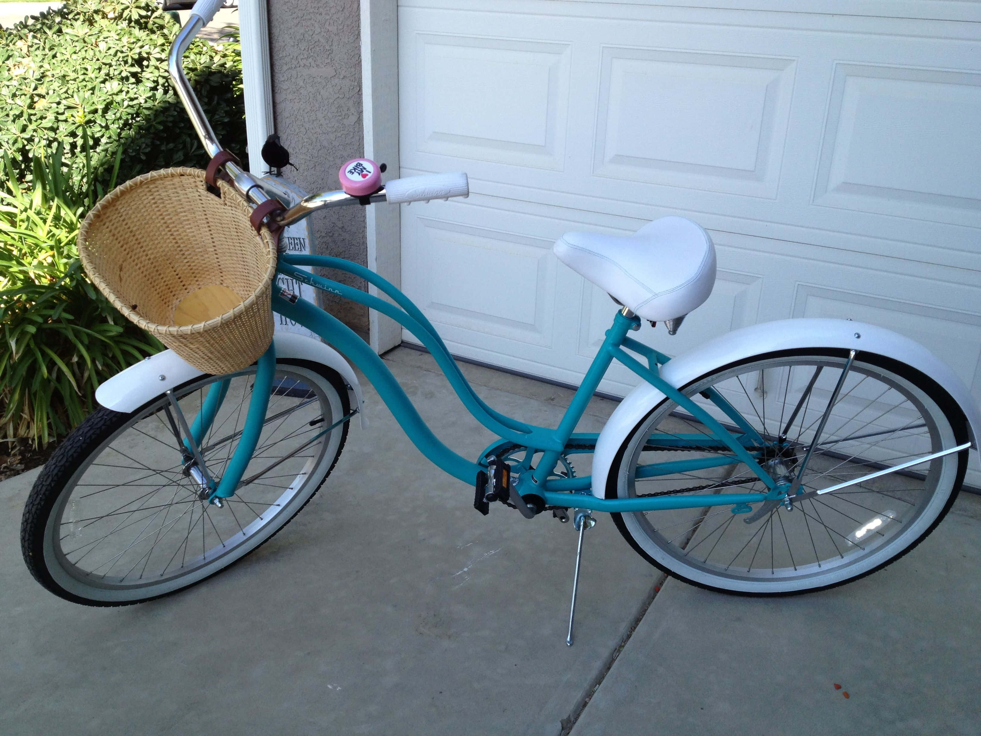Beach Cruiser 桌布 壁紙 — 一輛藍綠色沙灘巡洋艦自行車，配有白色擋泥板和柳條籃，停在白色車庫門旁的混凝土車道上。