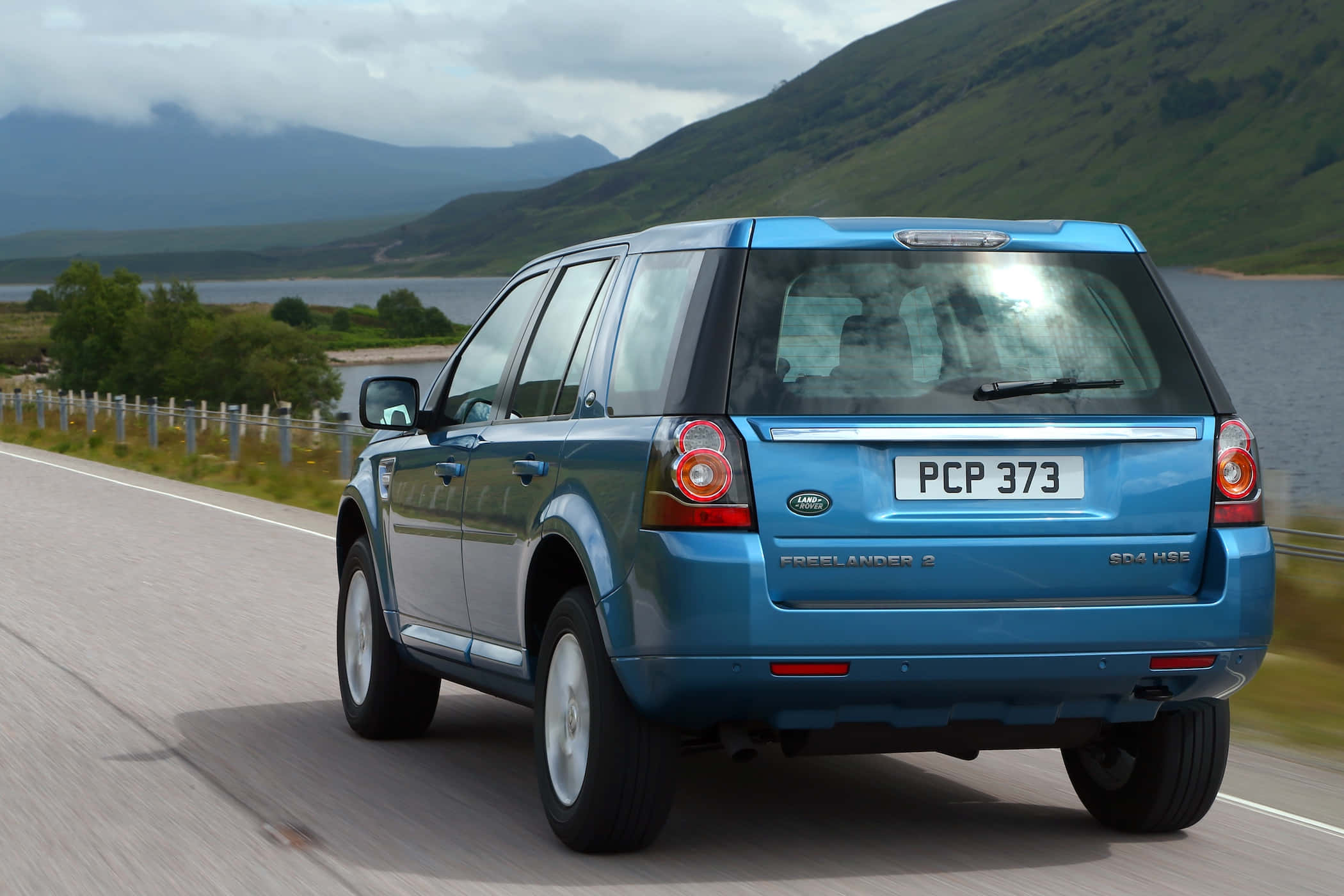 Land Rover Freelander 桌布 壁紙 — 一輛藍色 Land Rover Freelander 2 沿著湖邊蜿蜒的道路行駛，背景是霧氣繚繞的山脈。
