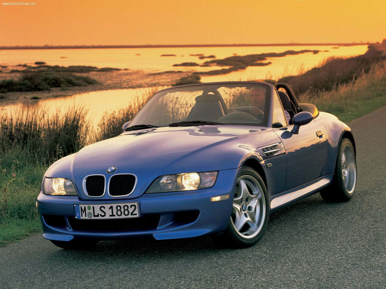 Bmw Z3 桌布 壁紙 — 一輛藍色BMW Z3開篷跑車在金色日落時分，背景有沼澤地的海岸公路上閃閃發光。