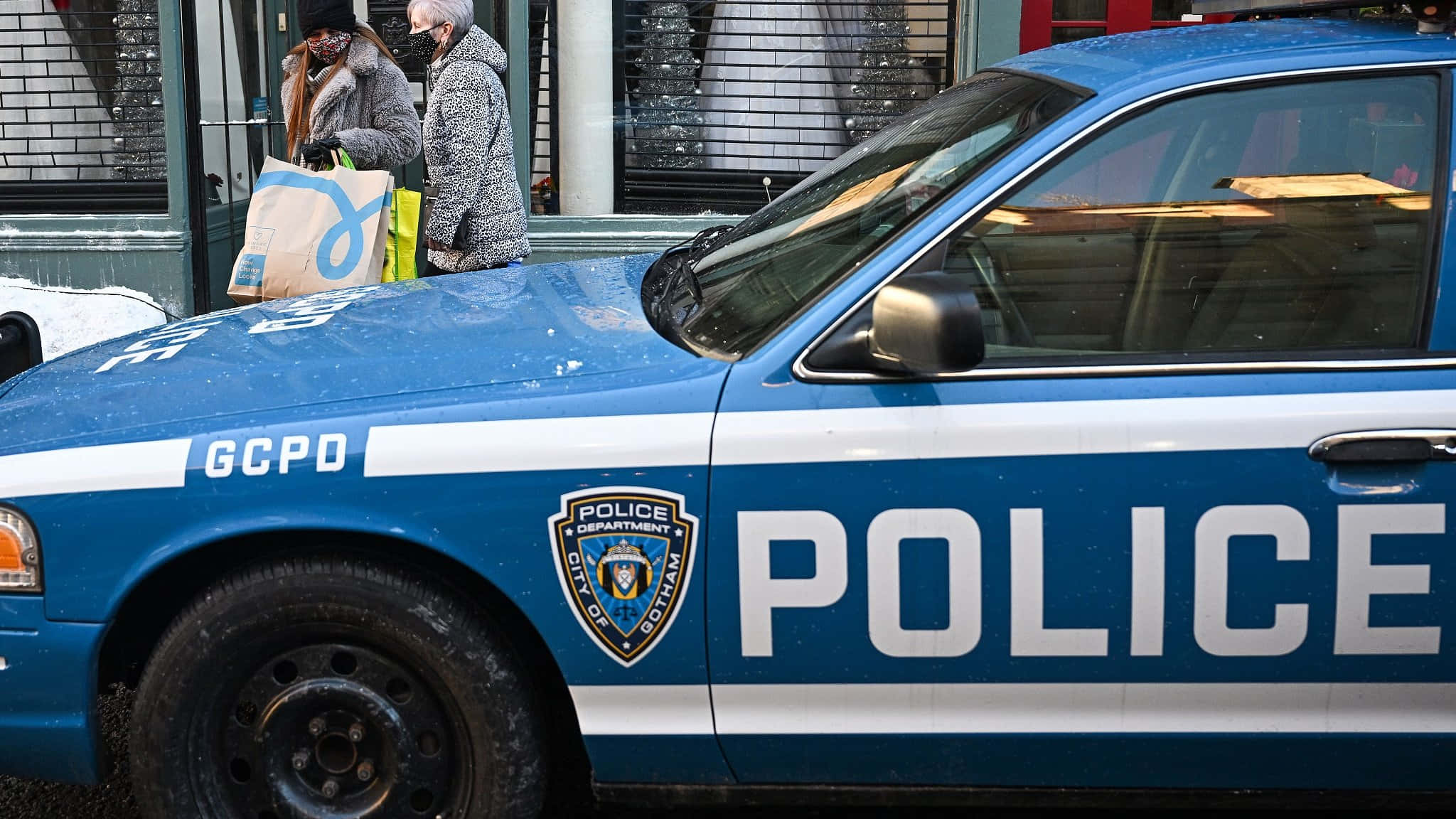 Gotham City Police Department 桌布 壁紙 — 一輛藍色GCPD警車停泊在積雪的街道上，兩名行人手持購物袋走過。