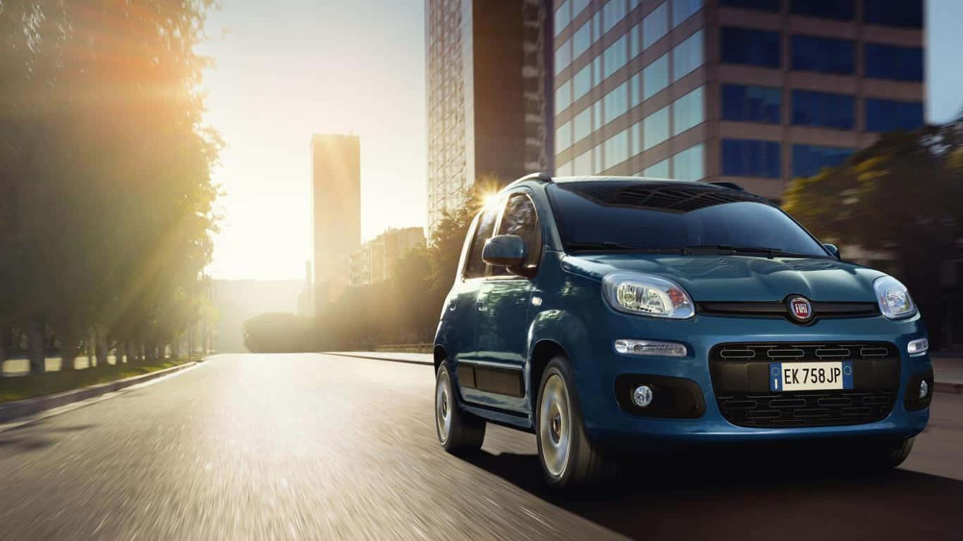 Fiat Panda 桌布 壁紙 — 一輛藍色嘅Fiat Panda喺陽光普照嘅城市街道上行駛，兩旁有現代建築物同樹木。