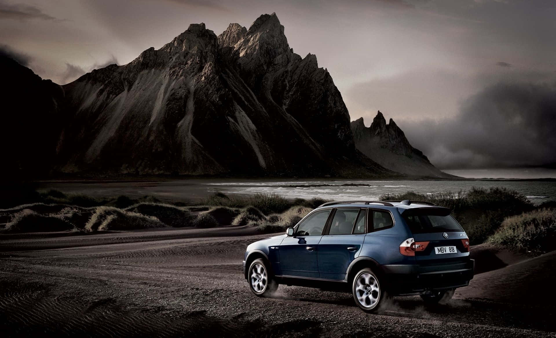 Bmw X3 桌布 壁紙 — 一輛藍色寶馬SUV在戲劇性的黑暗山脈和暴風雨天空下行駛在碎石路上。