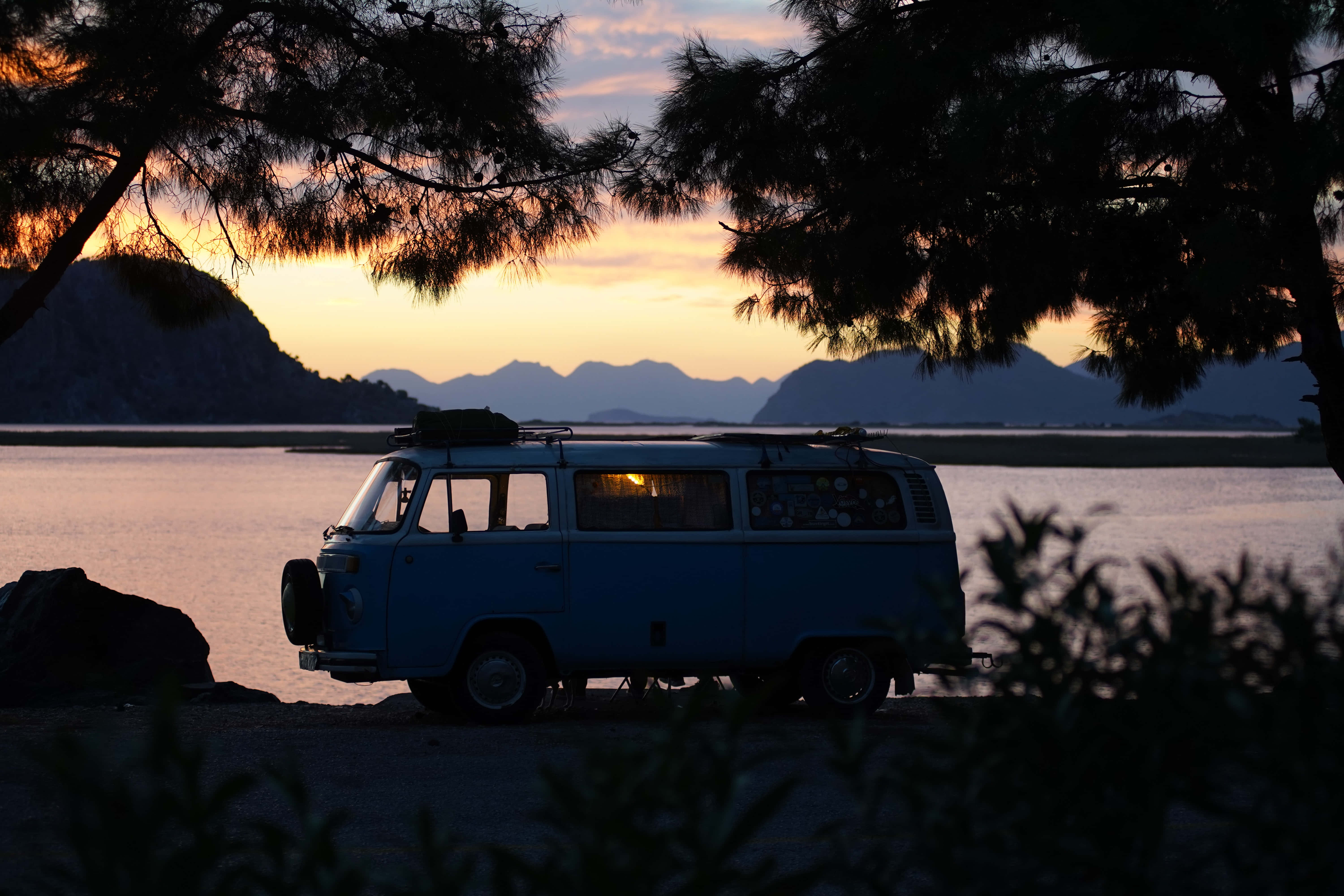 road trip 桌布 壁紙 — 一輛藍色復古麵包車停在海灘上，夕陽西下，背景是群山，被樹枝框住。
