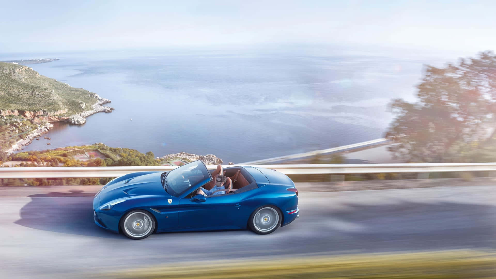 Ferrari California T 桌布 壁紙 — 一輛藍色法拉利敞篷車沿著海岸公路疾馳，司機揮手致意，俯瞰廣闊的海洋和陽光照耀的懸崖。