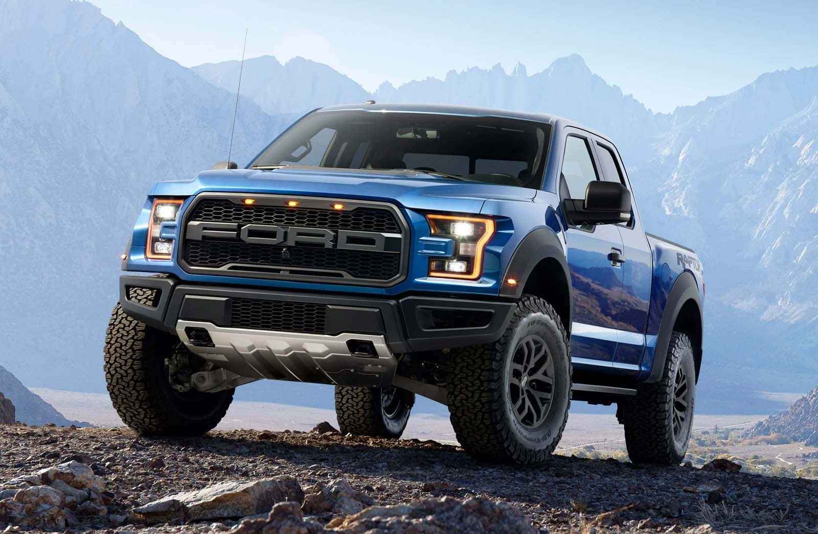 Ford Ranger 桌布 壁紙 — 一輛藍色福特 F-150 Raptor 卡車配備大型輪胎，在晴朗的天空下，以遠處積雪的山峰為背景，停在岩石地形上。
