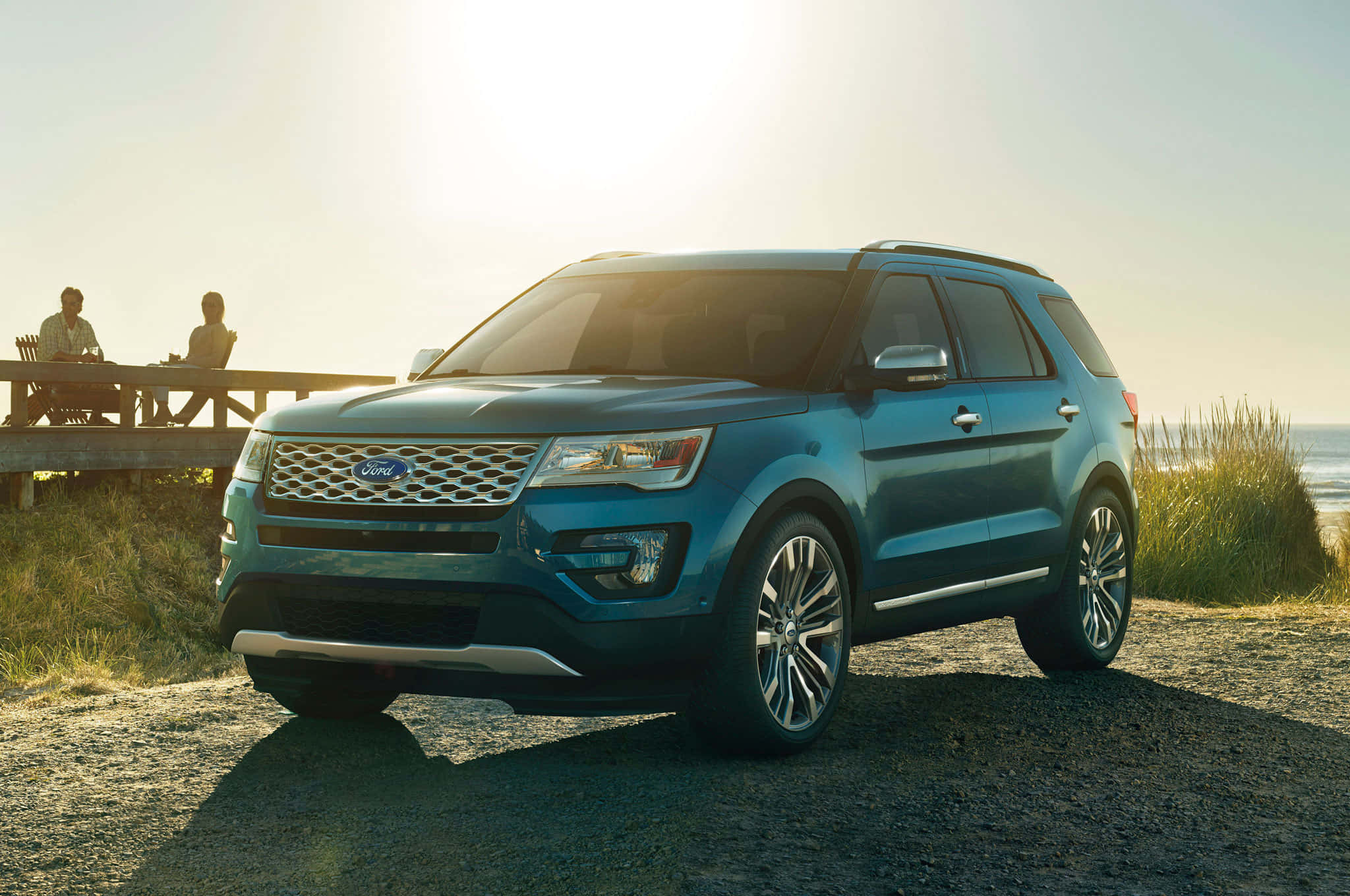 Ford Explorer 桌布 壁紙 — 一輛藍色福特Explorer SUV停在夕陽下的礫石海灘上，背景中有兩人坐在木桌旁。