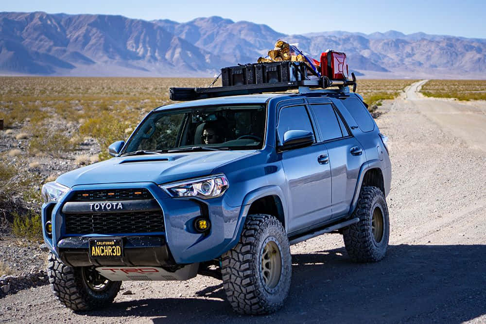 Off-road Car 桌布 壁紙 — 一輛藍色豐田4Runner配備超大輪胎和車頂裝備，在沙漠碎石路上行駛，背景是山脈。