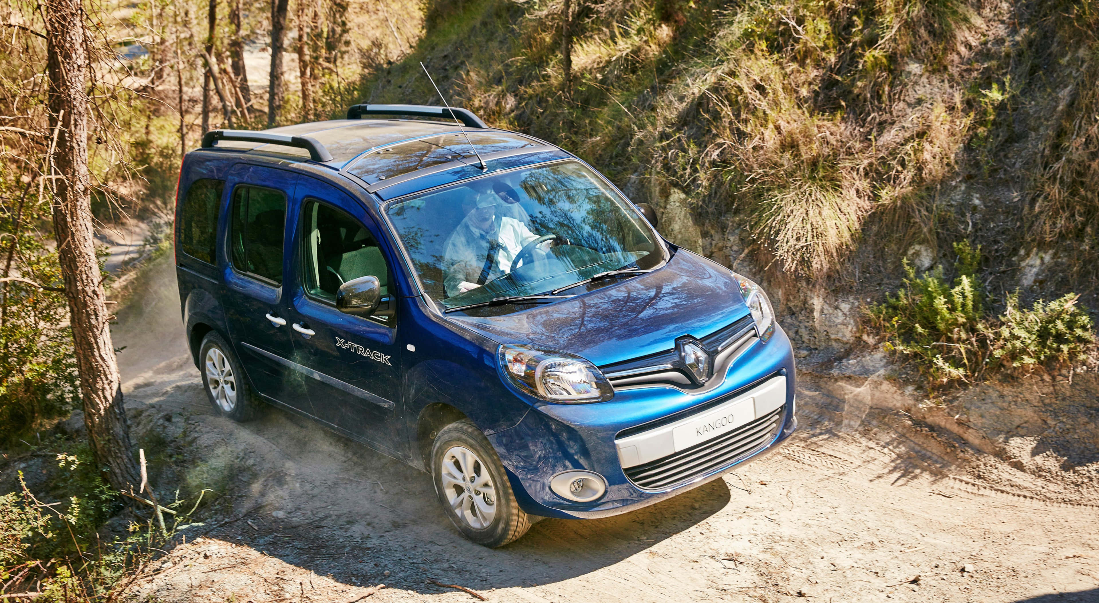Renault Kangoo 桌布 壁紙 — 一輛藍色雷諾Kangoo Trekking在塵土飛揚的森林小徑上行駛，周圍環繞著高大的樹木和岩石地形。