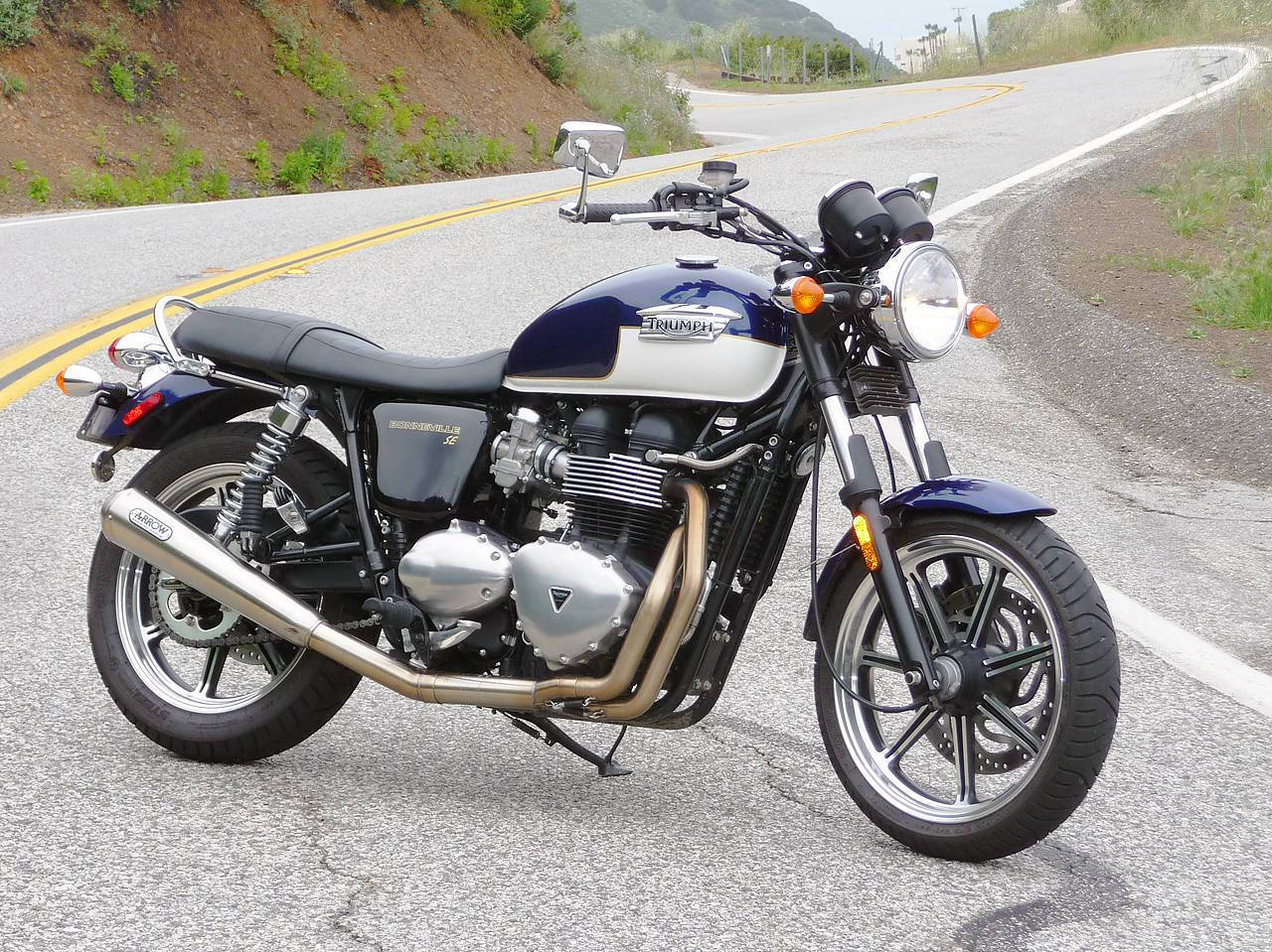 Triumph Bonneville Blue SE 壁紙 — 一輛藍銀色的Triumph電單車停泊在蜿蜒的瀝青路上，背景有山丘和稀疏的綠植。
