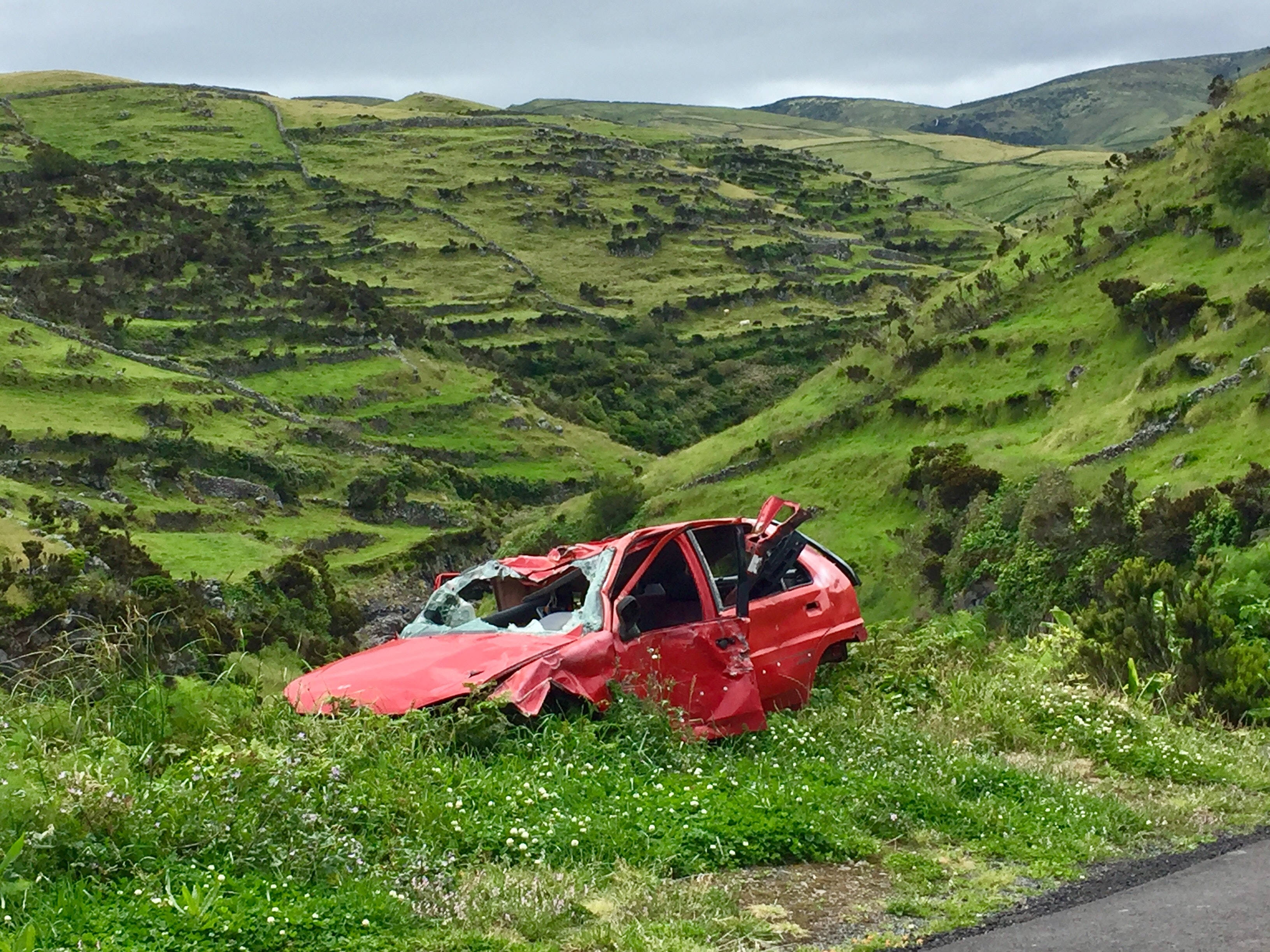 car crash 桌布 壁紙 — 一輛被壓碎的紅色汽車被遺棄在路邊的草溝裡，周圍是起伏的綠色山丘，天空多雲。