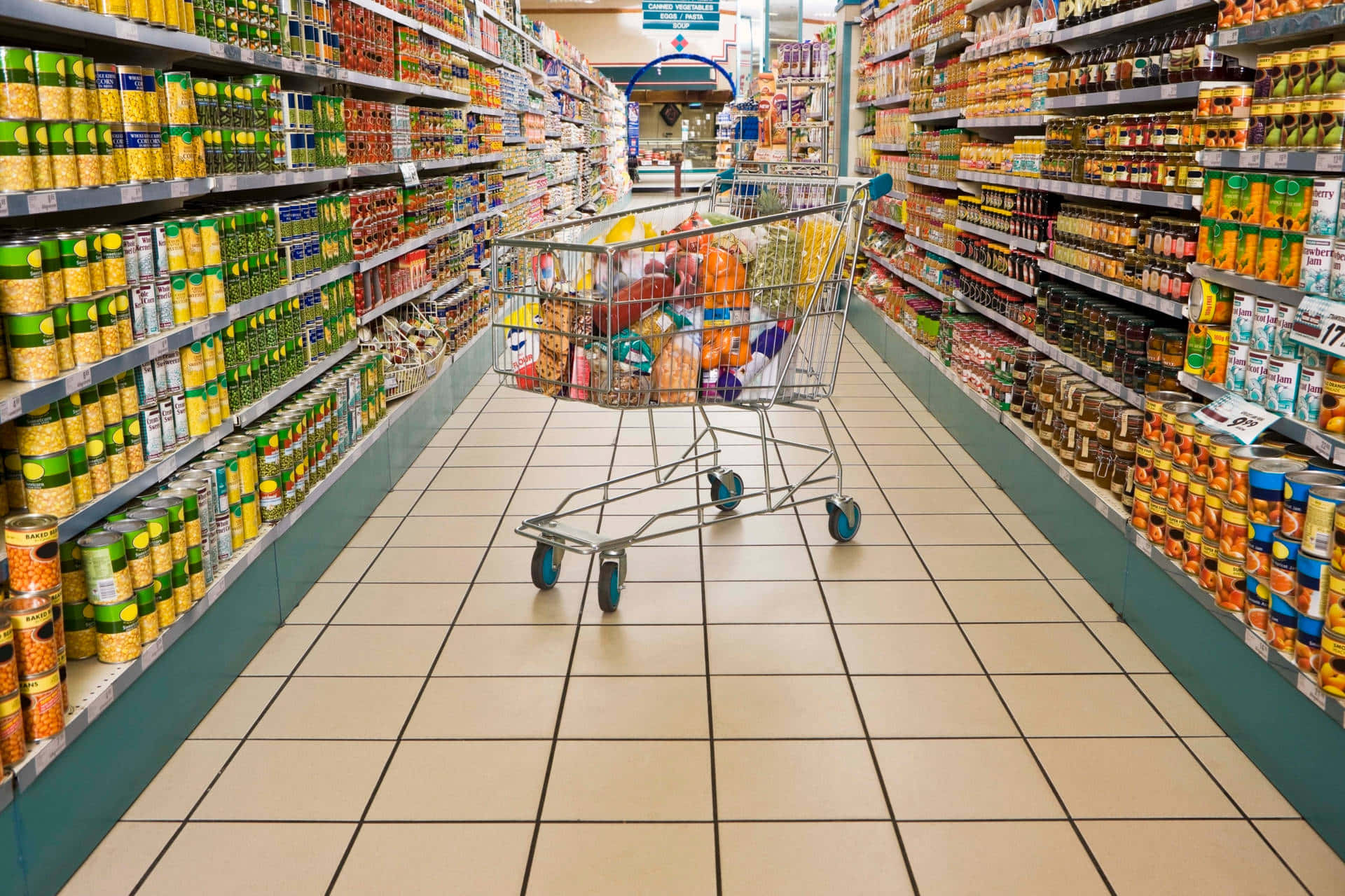 Grocery Store 桌布 壁紙 — 一輛裝滿雜貨的購物車放在光線明亮的超市通道中央，通道兩旁貨架陳列整齊。