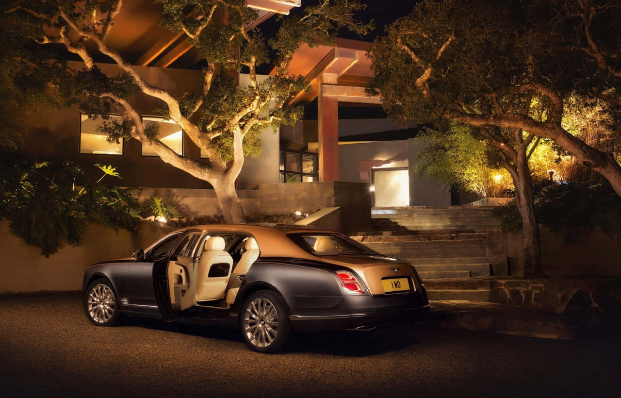 Bentley Mulsanne 桌布 壁紙 — 一輛豪華賓利房車打開車門，在溫暖的夜燈下閃閃發光，旁邊是一座被綠樹環繞的現代住宅。