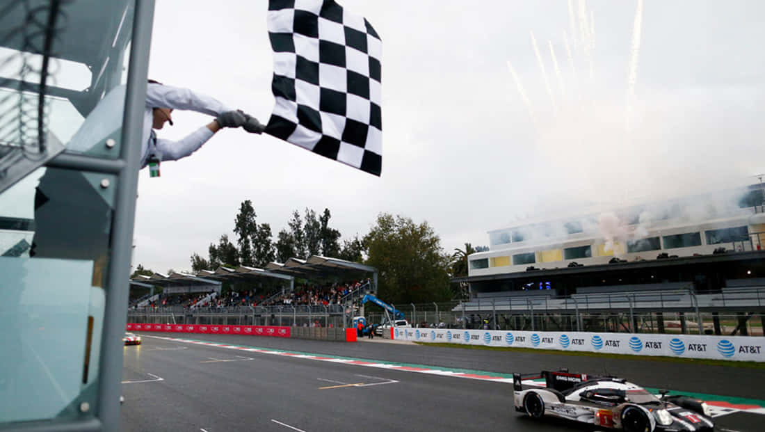 Chequered Flag 桌布 壁紙 — 一輛賽車越過終點線，旗手揮舞著格子旗，伴隨著慶祝的煙火和多雲的天空。