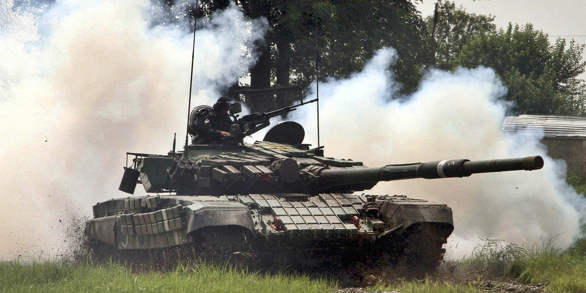 Indian Army 桌布 壁紙 — 一輛軍用坦克在草地上發射大炮，揚起煙霧和泥土，一名士兵操作炮塔。