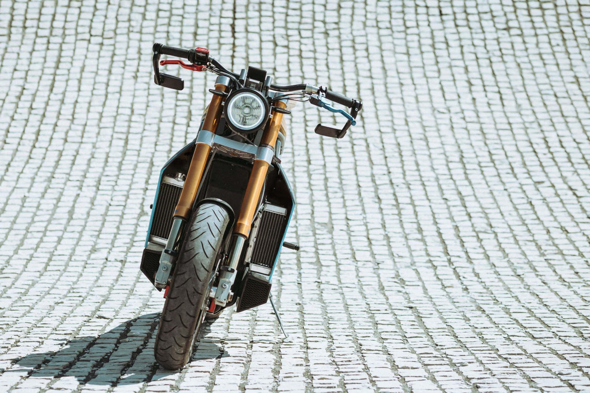Electric Bike 桌布 壁紙 — 一輛配備金色前叉和黑色飾件的訂製電單車，停泊在陽光普照的鵝卵石街道上，投下清晰的陰影。