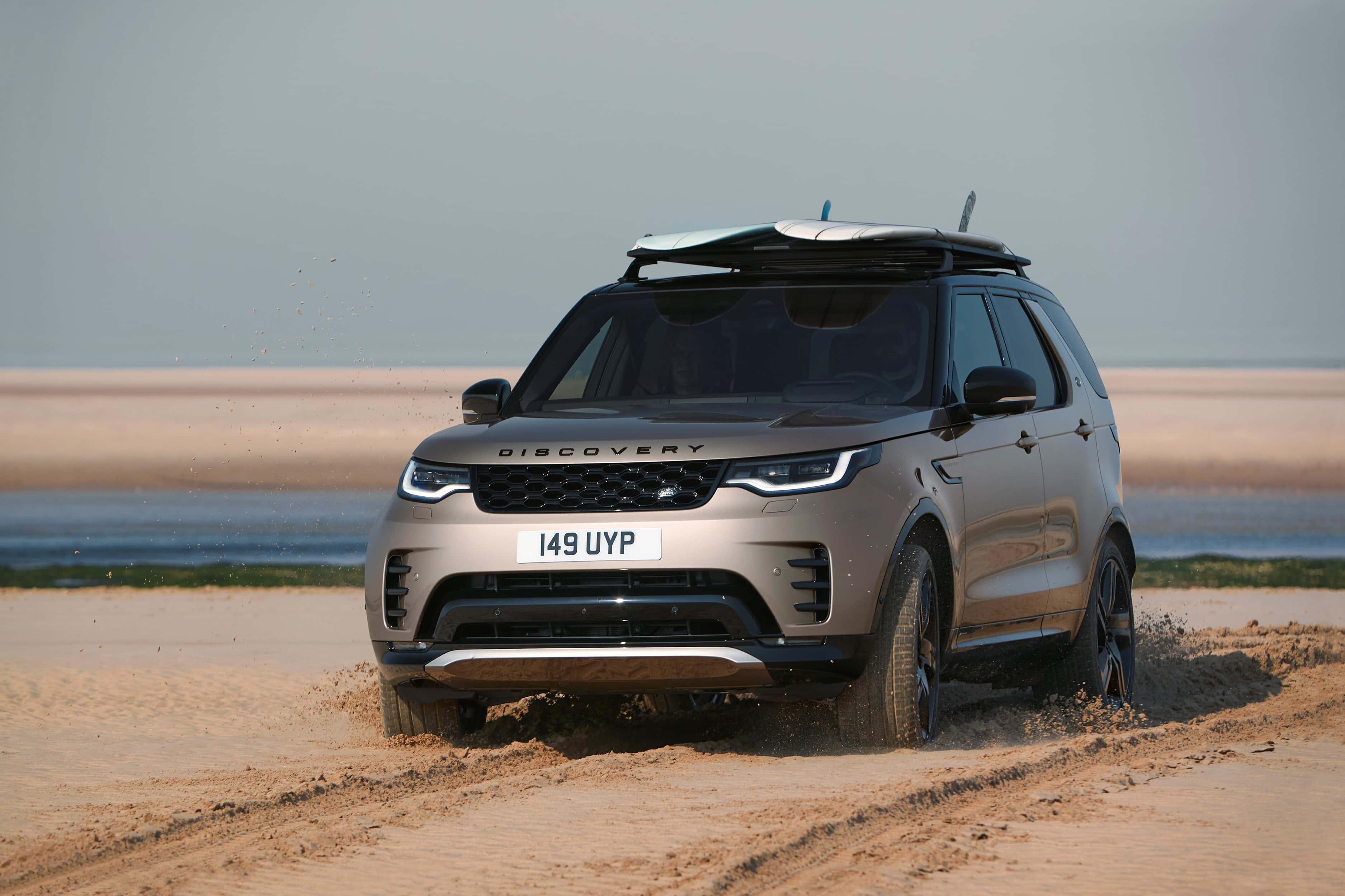 Land Rover Discovery 桌布 壁紙 — 一輛金屬色SUV車頂載著衝浪板，在陰天平靜的海邊濕沙上行駛。