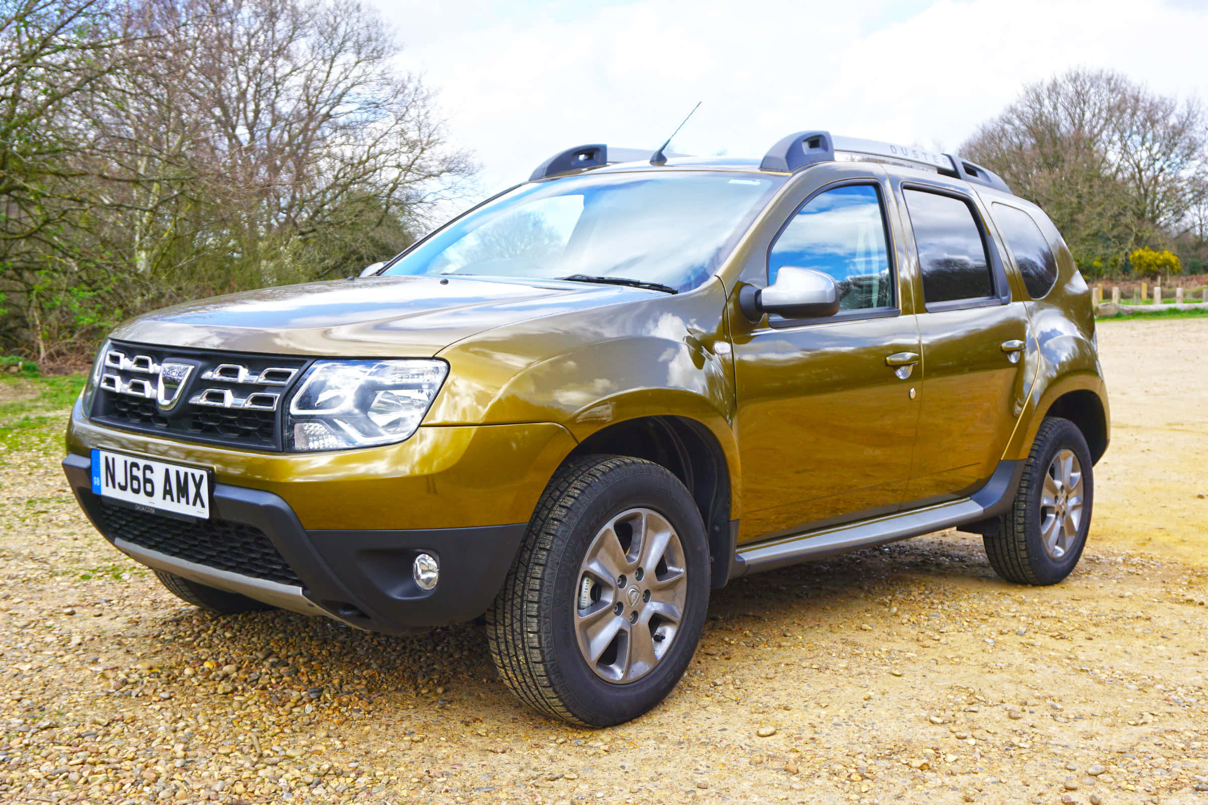 Dacia 桌布 壁紙 — 一輛金屬金色的Dacia Duster SUV停泊在碎石上，背景是光禿禿的樹木和籬笆，天空多雲。