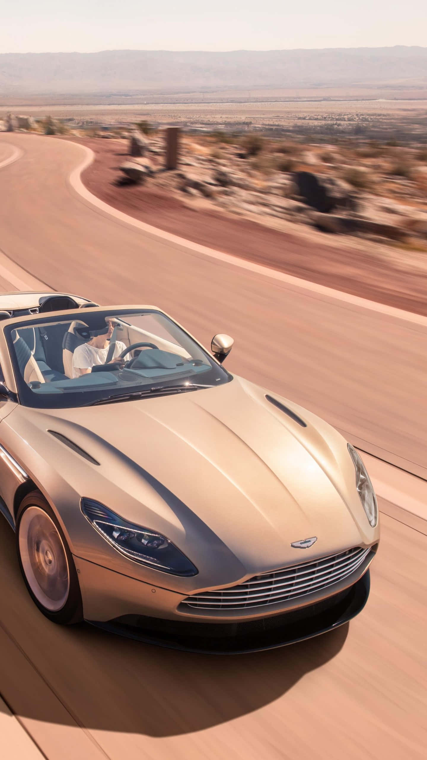 Aston Martin Db11 桌布 壁紙 — 一輛金色 Aston Martin DB11 Volante 在明亮的陽光下，沿著蜿蜒的沙漠公路疾馳，背景是模糊的風景和遠山。