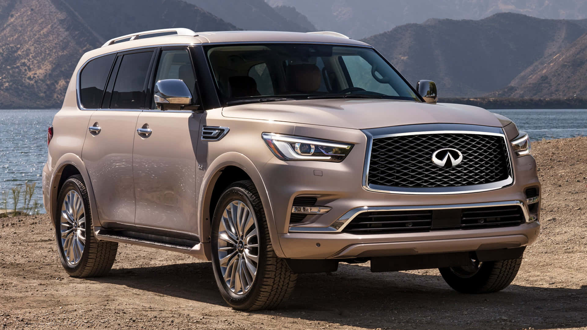 Infiniti Qx80 桌布 壁紙 — 一輛金色 Infiniti QX80 SUV 停泊在沙灘上，旁邊是平靜的水面，背景有山巒。