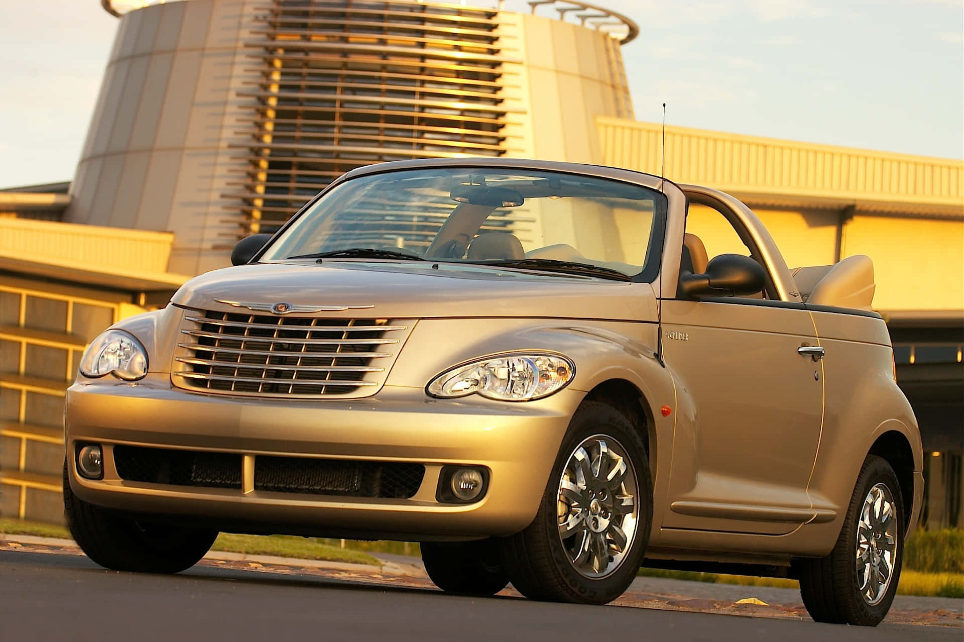 Chrysler Pt Cruiser 桌布 壁紙 — 一輛金色克萊斯勒 PT Cruiser 敞篷車停放在戶外，背景是現代建築，沐浴在溫暖的金色光線下。