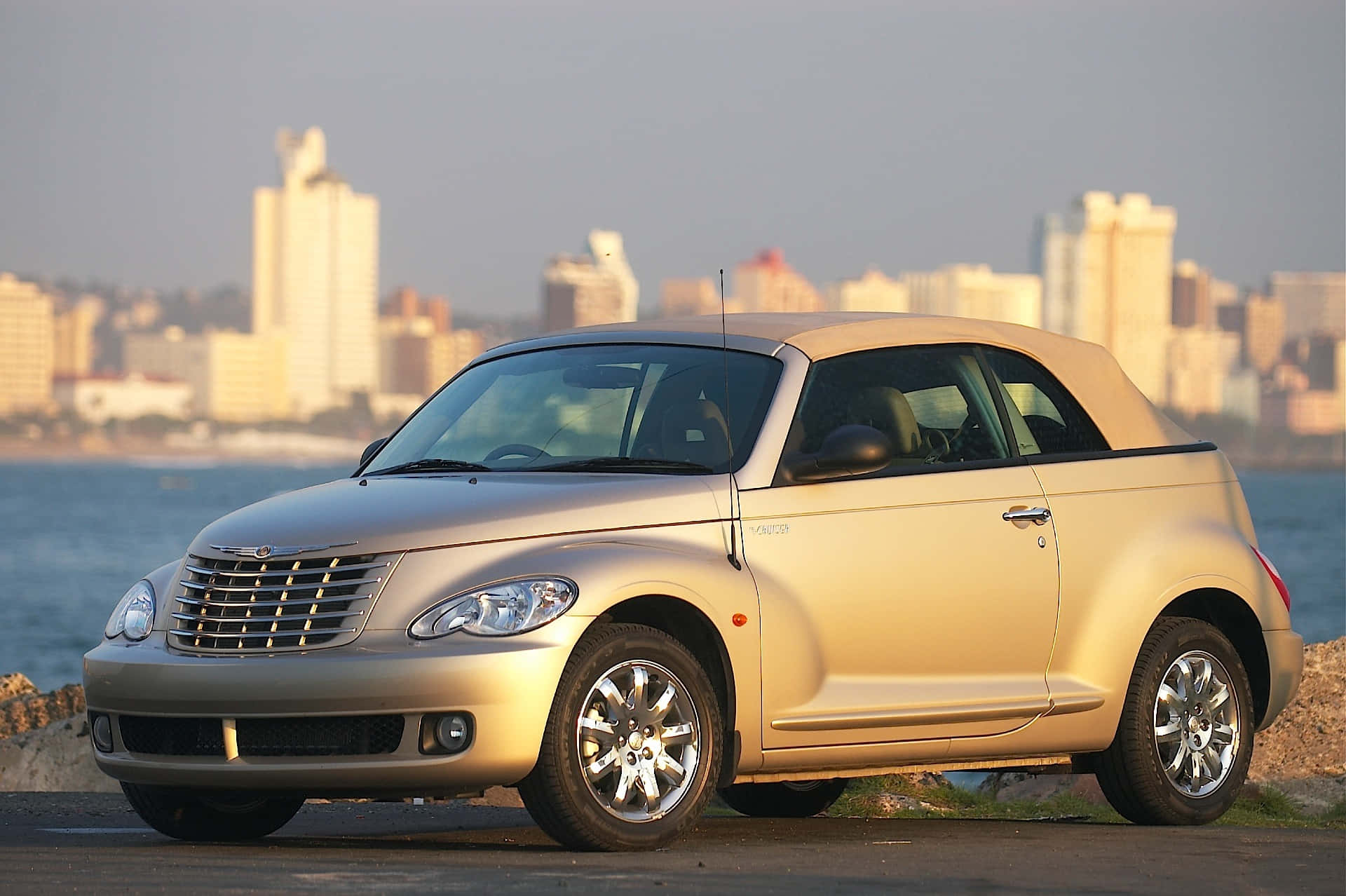 Chrysler Pt Cruiser 桌布 壁紙 — 一輛金色克萊斯勒 PT Cruiser 敞篷車停在水邊，背景是日落時分的城市天際線。
