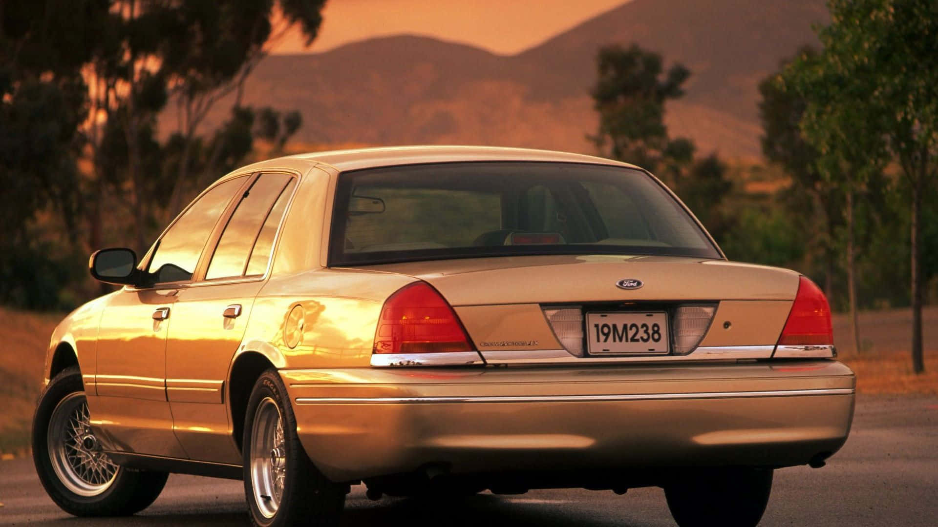 Ford Crown Victoria 桌布 壁紙 — 一輛金色福特皇冠維多利亞轎車在溫暖的日落光線下閃閃發光，停在路邊，背景有山脈和樹木。