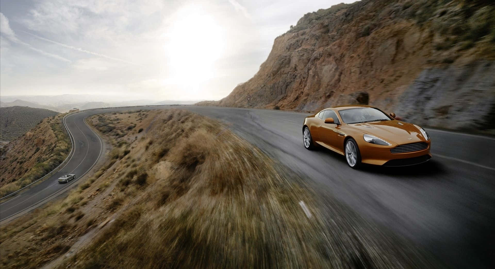 Aston Martin Db9 桌布 壁紙 — 一輛金色跑車在明亮的天空下，沿著蜿蜒的山路疾馳，帶有模糊的動感和岩石地形。