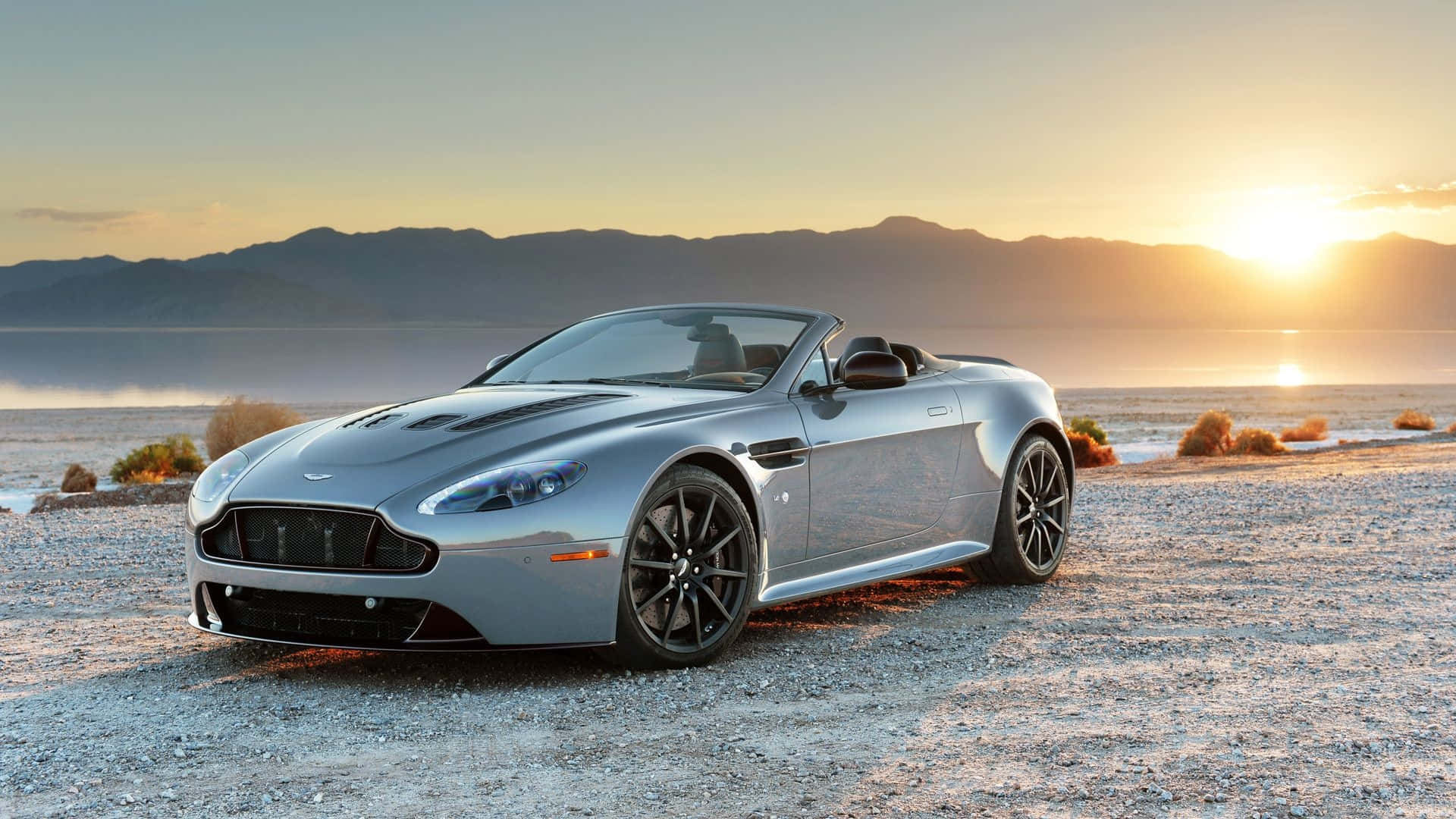 Aston Martin V12 Vantage 桌布 壁紙 — 一輛銀色 Aston Martin V12 Vantage 開篷車停在日落時分的碎石沙漠海岸上，背景有山脈。