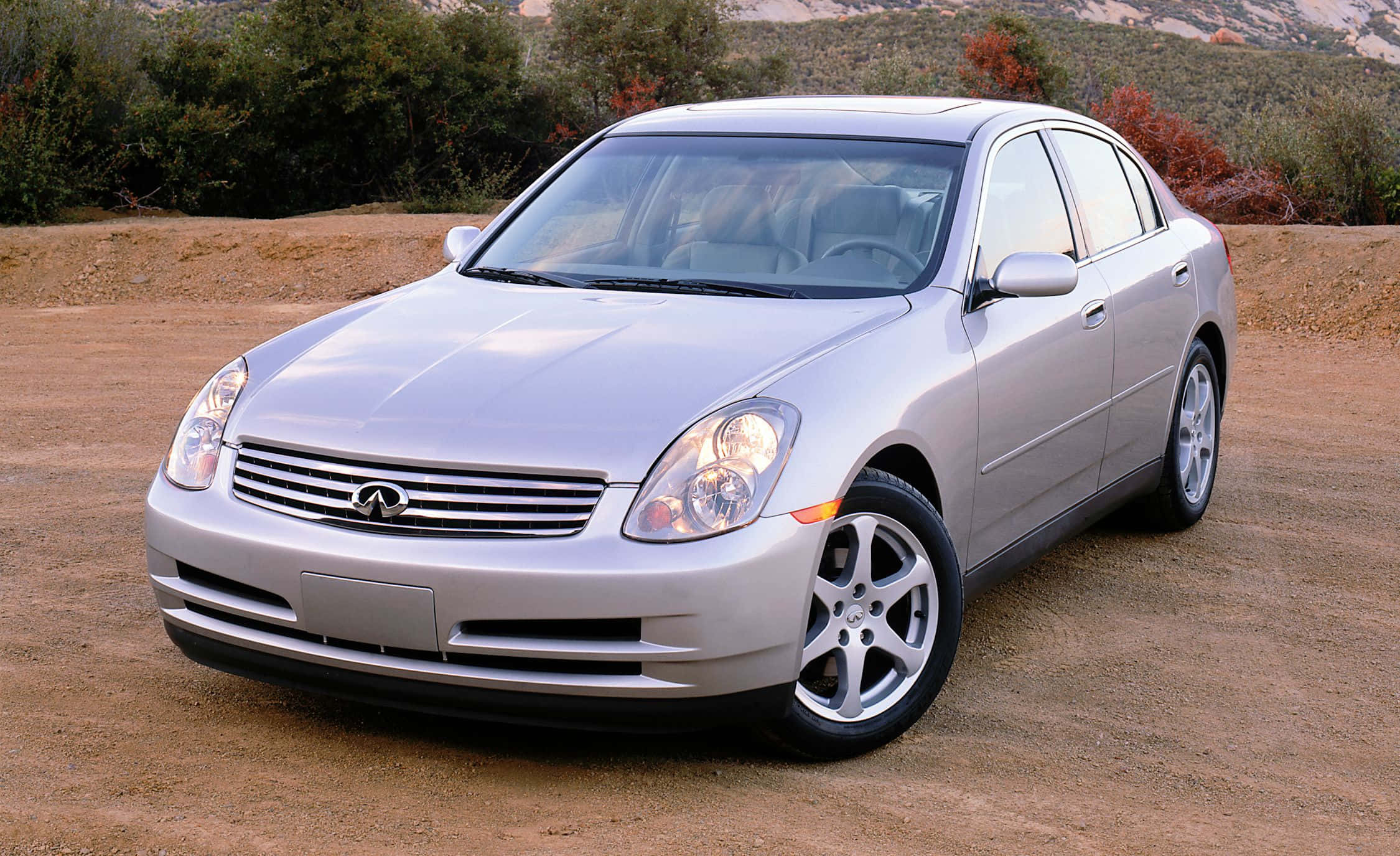 Infiniti G35 桌布 壁紙 — 一輛銀色 Infiniti 房車停泊在泥路上，背景有灌木叢和山丘。