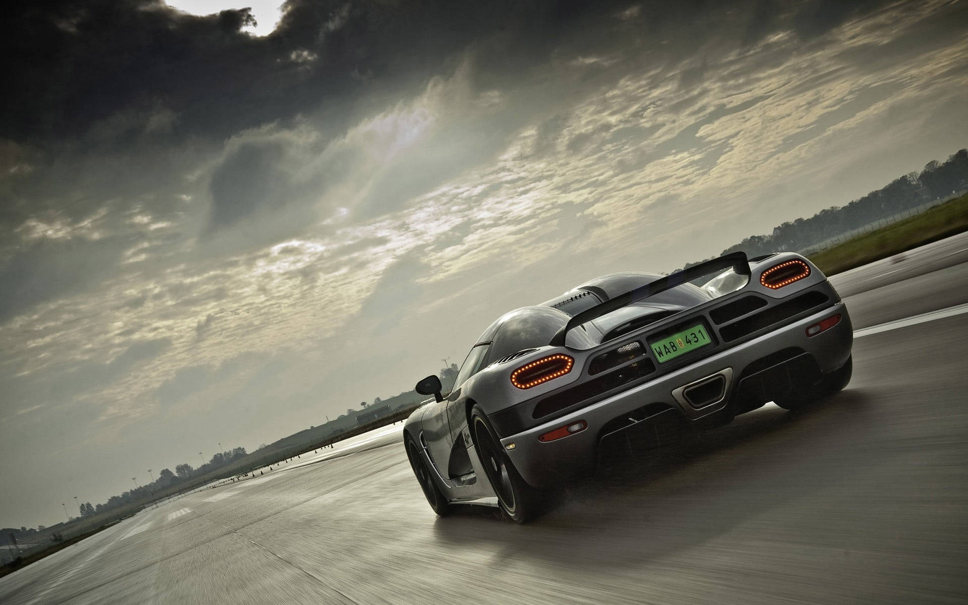 Sports Car 桌布 壁紙 — 一輛銀色 Koenigsegg 超級跑車在潮濕的賽道上疾馳，天空烏雲密佈，陽光穿透雲層，場面壯觀。