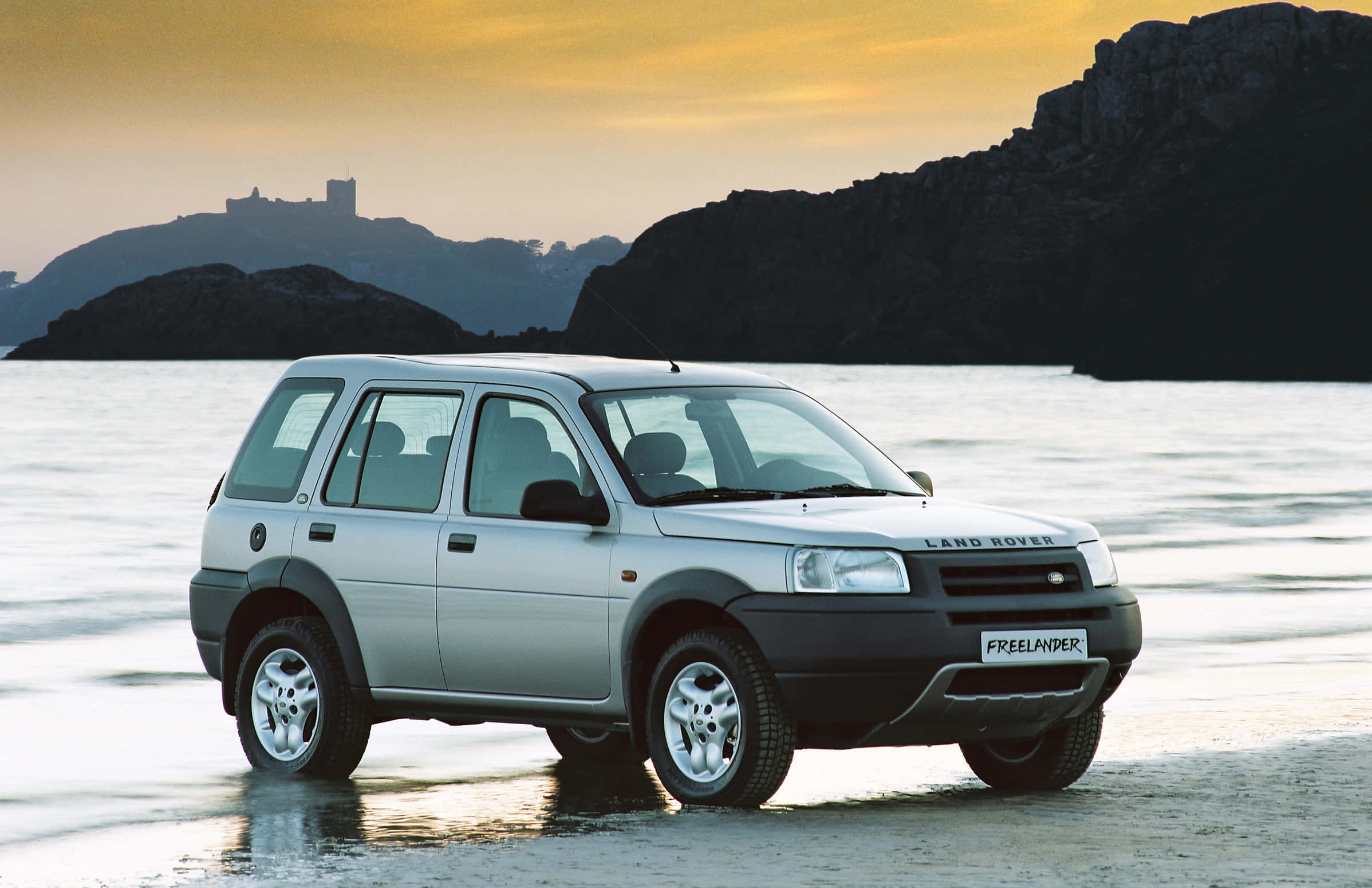 Land Rover Freelander 桌布 壁紙 — 一輛銀色 Land Rover Freelander 在日落時分停泊在濕漉漉的沙灘上，背景是岩石峭壁和遠處的城堡。