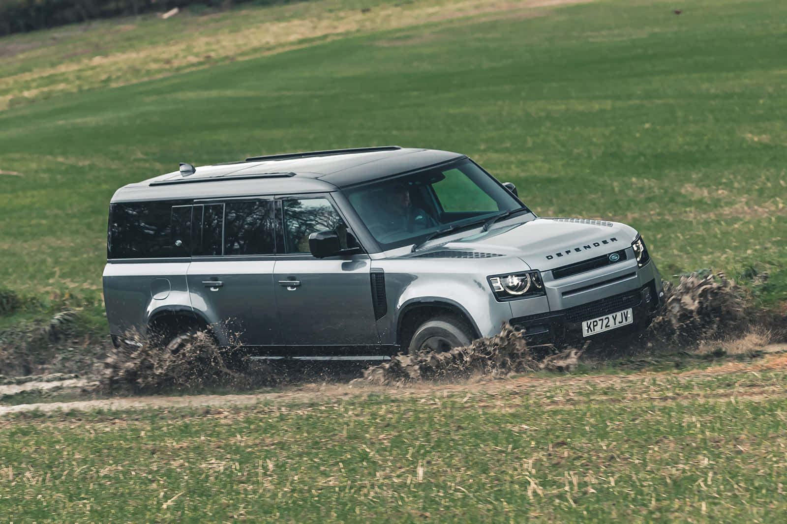 Land Rover Defender 桌布 壁紙 — 一輛銀色 Land Rover Defender 在泥濘地形上強勁行駛，在草地上濺起水花。