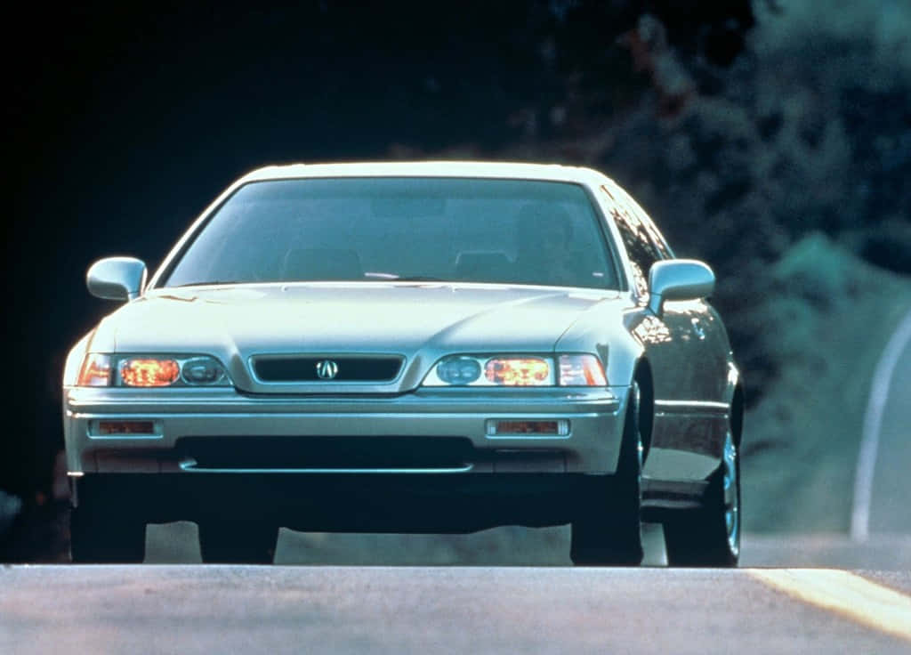 Acura Legend 桌布 壁紙 — 一輛銀色Acura房車亮著車頭燈，在柔和日光下沿著兩旁有暗樹的道路向前行駛。