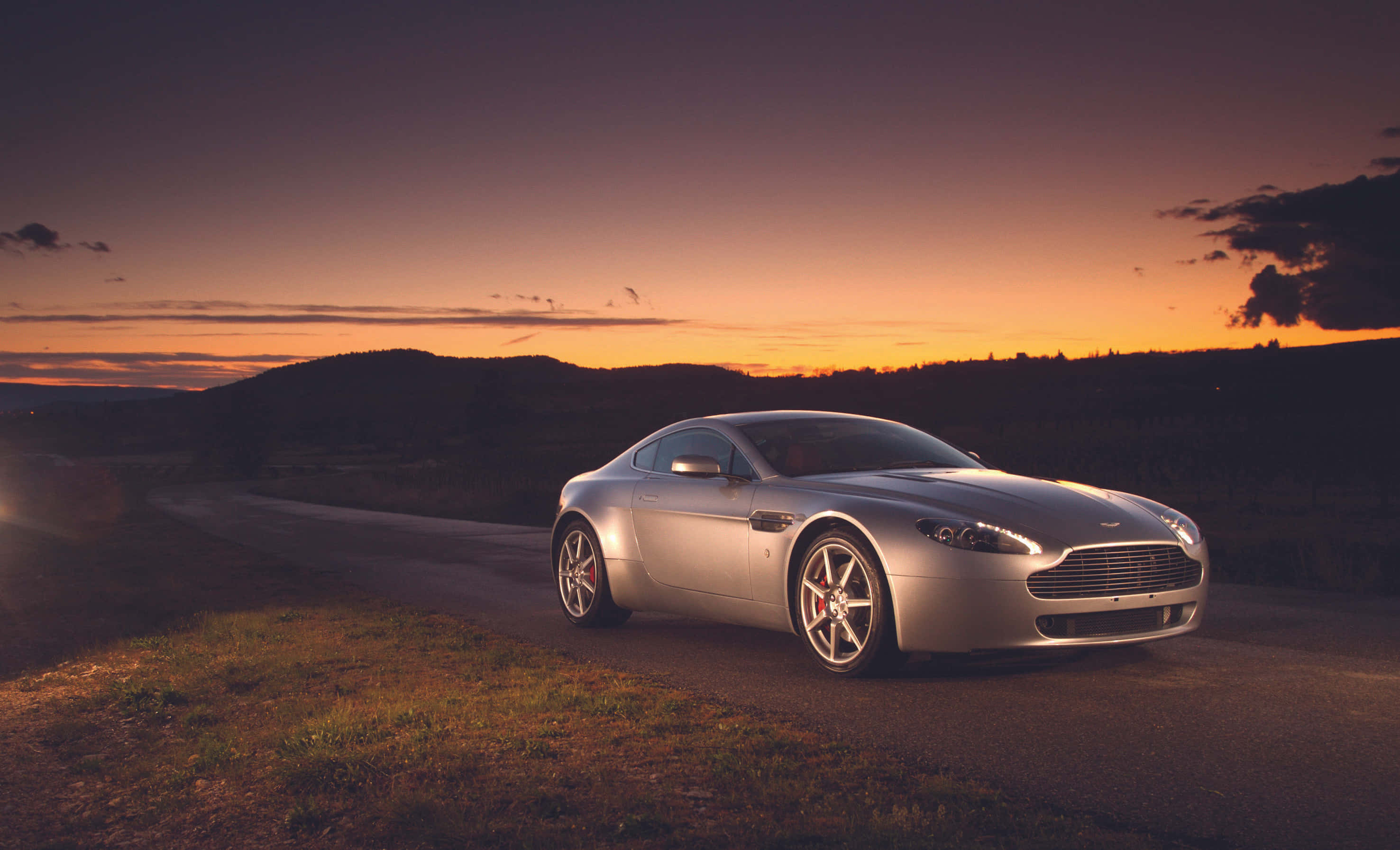 Aston Martin Vantage 桌布 壁紙 — 一輛銀色Aston Martin跑車在日落時分於蜿蜒道路上閃耀，背景是起伏的丘陵。