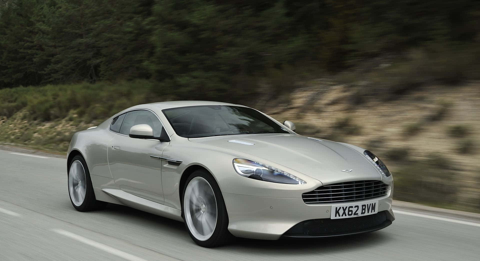 Aston Martin Db9 桌布 壁紙 — 一輛銀色Aston Martin跑車沿著蜿蜒的道路穿過樹木繁茂的山坡飛馳。