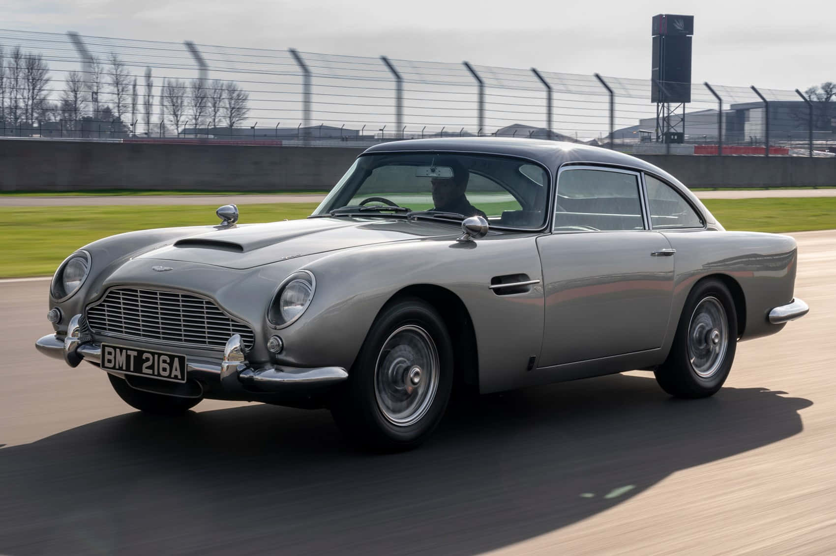 Aston Martin Db5 桌布 壁紙 — 一輛銀色Aston Martin DB5在賽道上疾馳，動作模糊，透過擋風玻璃可見司機。