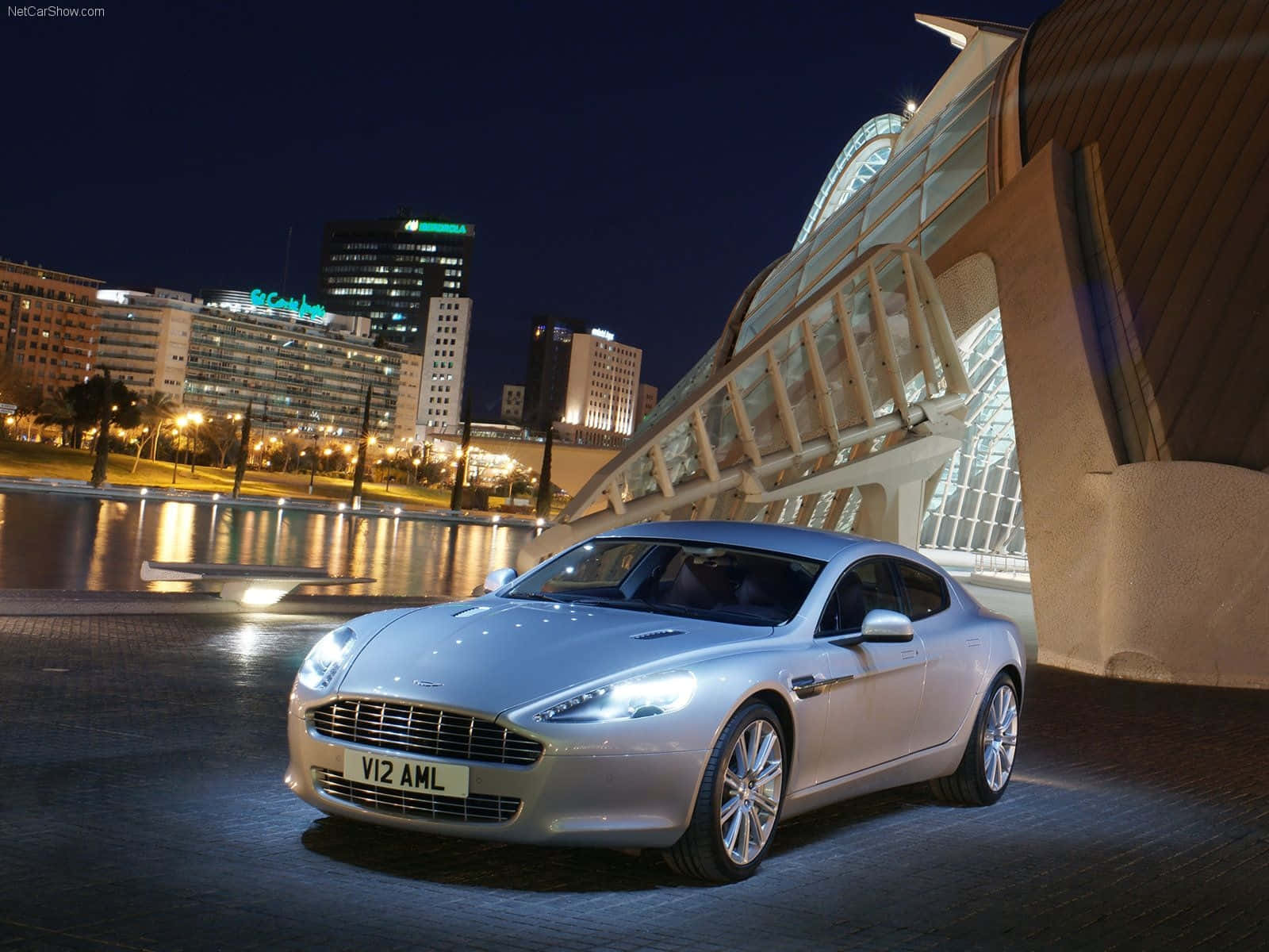 Aston Martin Rapide S 桌布 壁紙 — 一輛銀色Aston Martin跑車在夜燈下閃閃發光，旁邊是一座現代玻璃橋，背後的城市天際線倒映在水中。