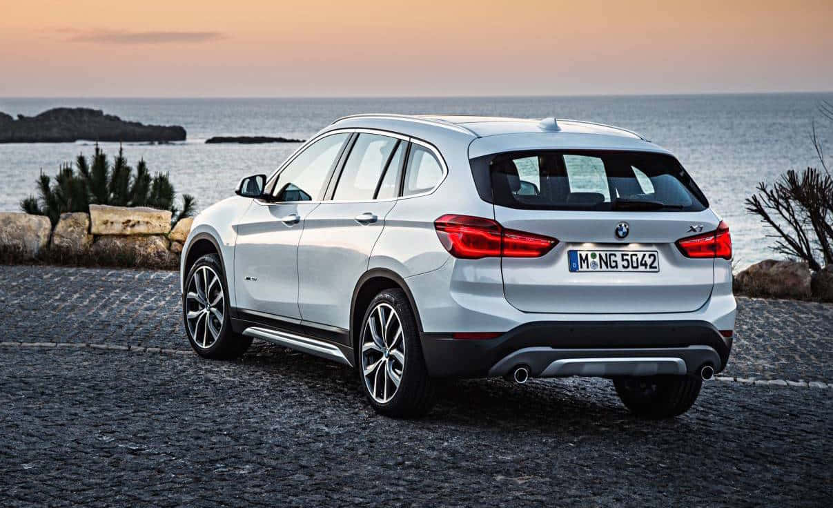 Bmw X1 桌布 壁紙 — 一輛銀色BMW X1 SUV停泊在日落時分海邊的鵝卵石小徑上，天空色調溫暖。