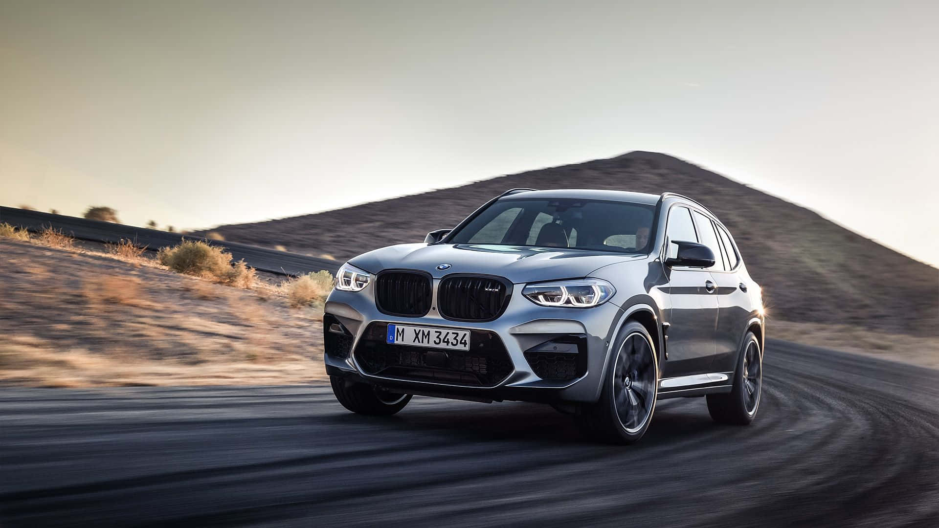 Bmw X3 桌布 壁紙 — 一輛銀色BMW X3 M SUV在蜿蜒的沙漠公路上高速行駛，背景是朦朧的山脈，呈現模糊的動態效果。