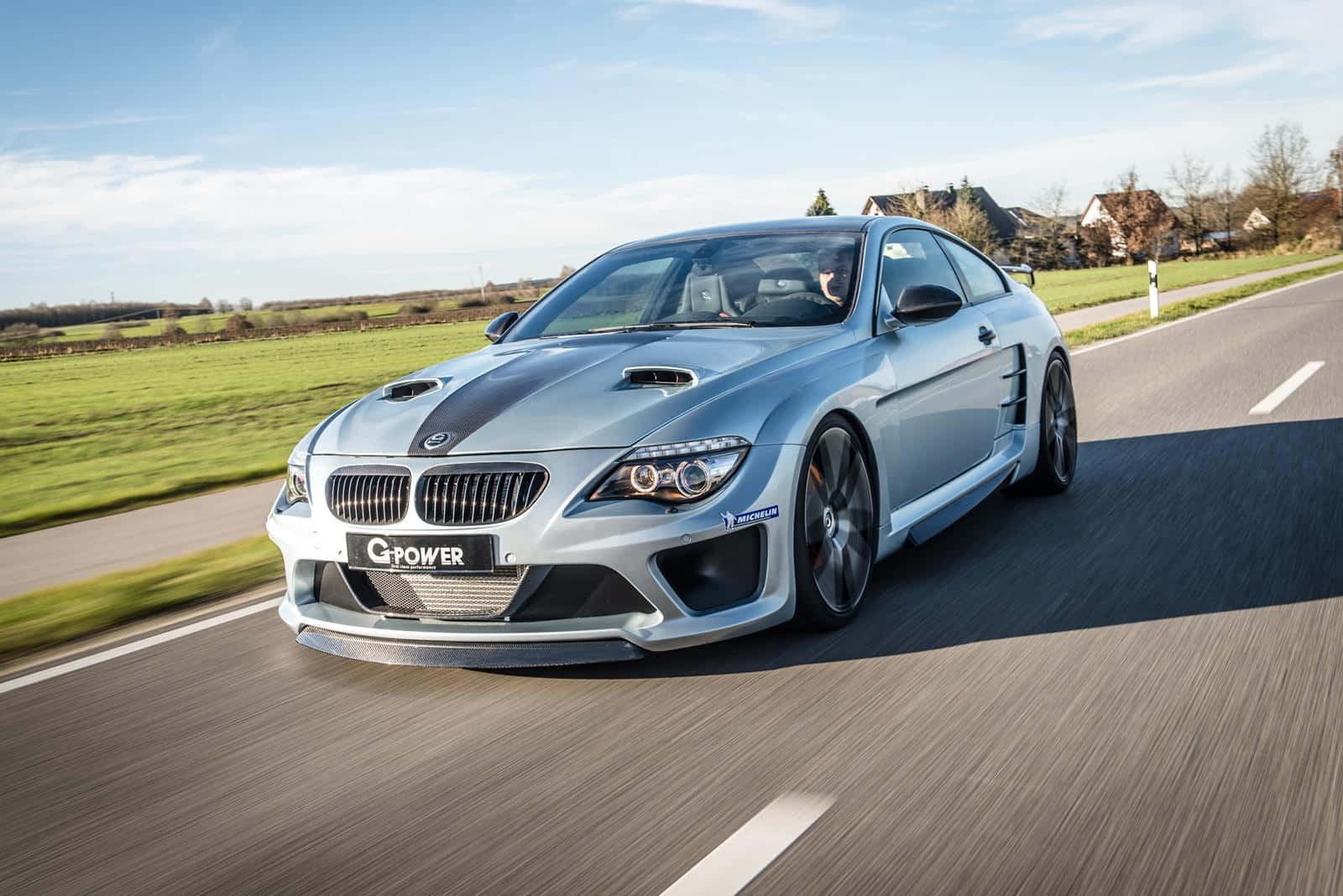 Bmw M6 桌布 壁紙 — 一輛銀色G-Power BMW M6跑車在鄉村道路上疾馳，背景是綠色的田野和房屋，天空清澈湛藍。