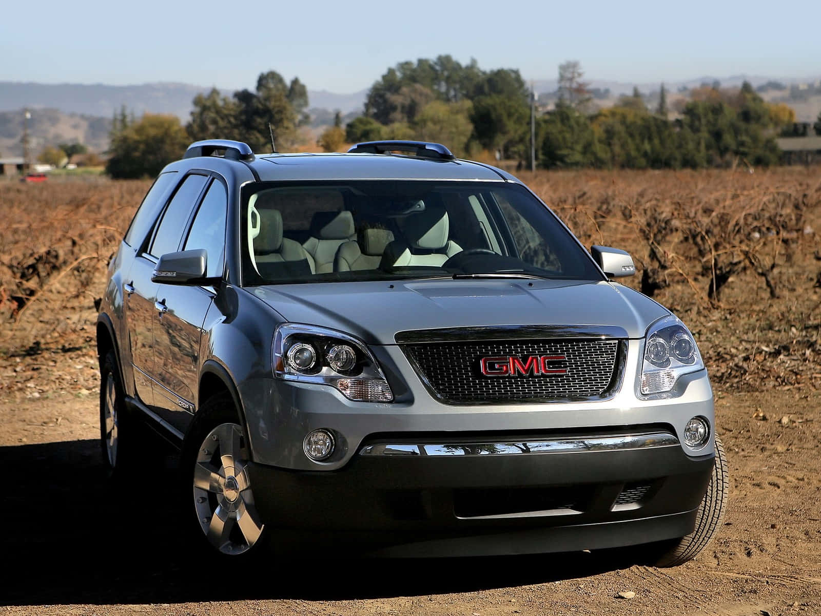 Gmc Acadia 桌布 壁紙 — 一輛銀色GMC Acadia SUV停泊在泥路上，旁邊有乾旱的葡萄園行和遠處的山丘，天空清澈湛藍。