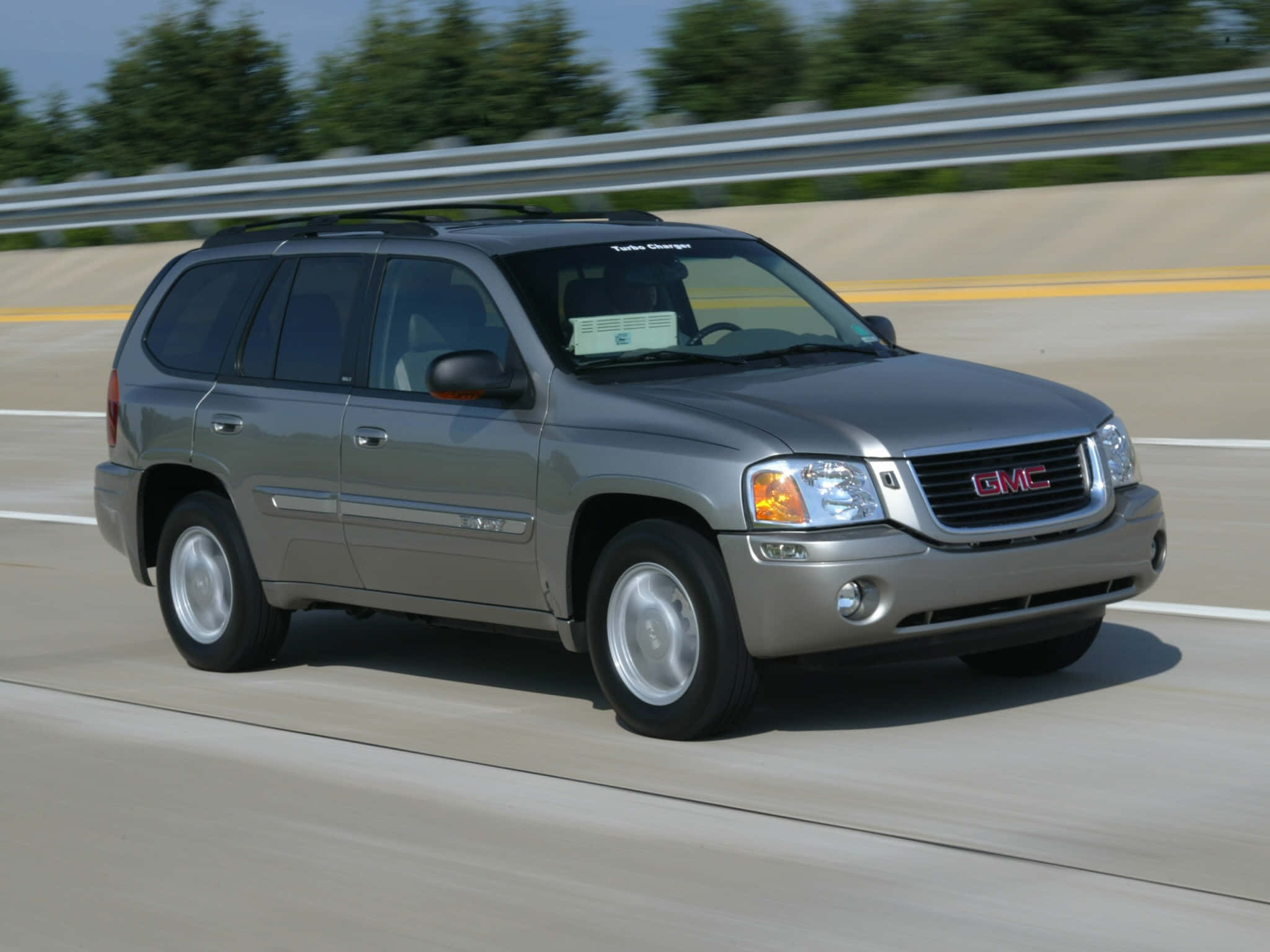 Gmc Envoy 桌布 壁紙 — 一輛銀色GMC Envoy SUV在陽光明媚的高速公路上疾馳，背景是模糊的綠樹和護欄。