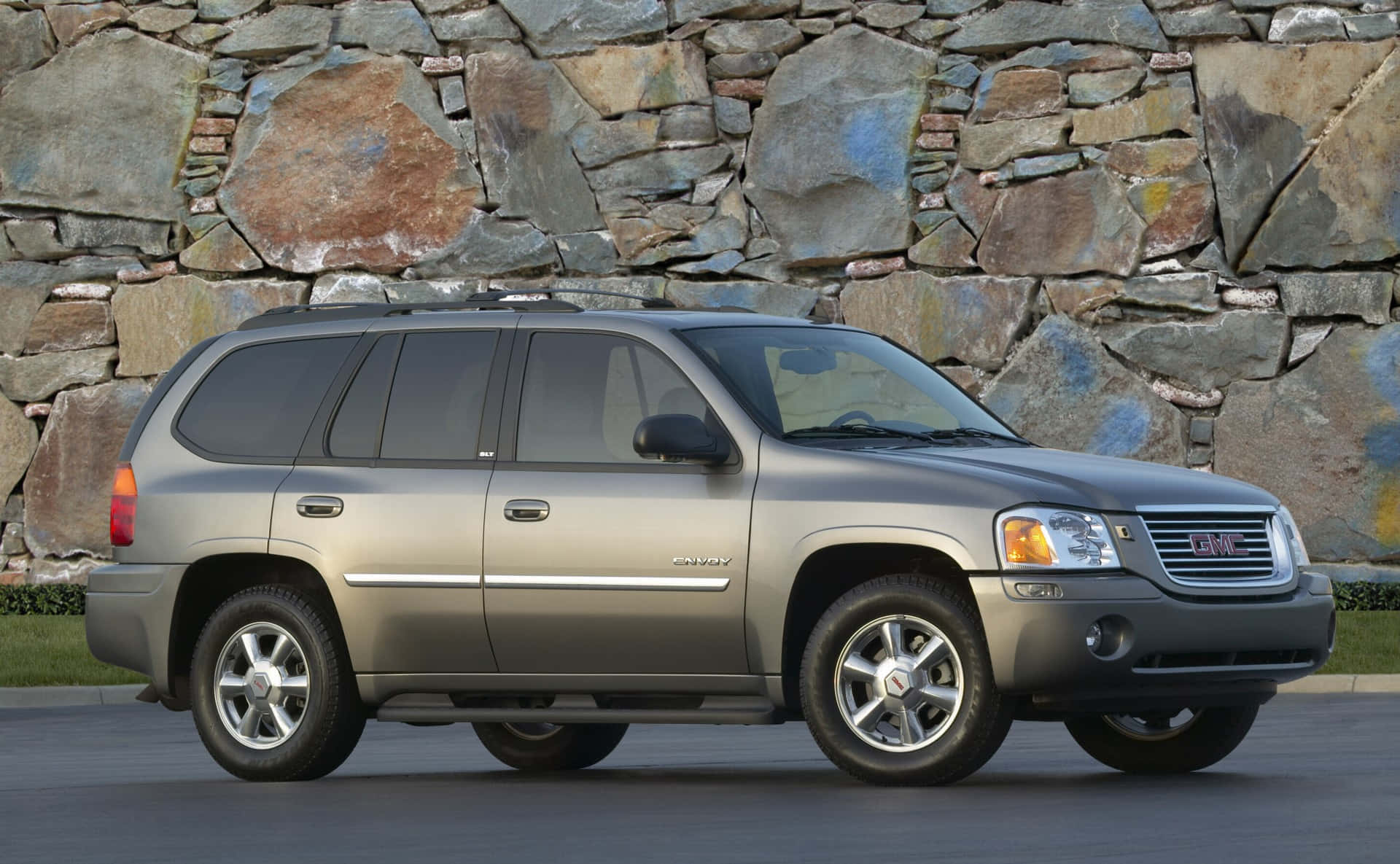 Gmc Envoy 桌布 壁紙 — 一輛銀色GMC Envoy SUV在明亮的日光下停放在粗獷的石牆前。