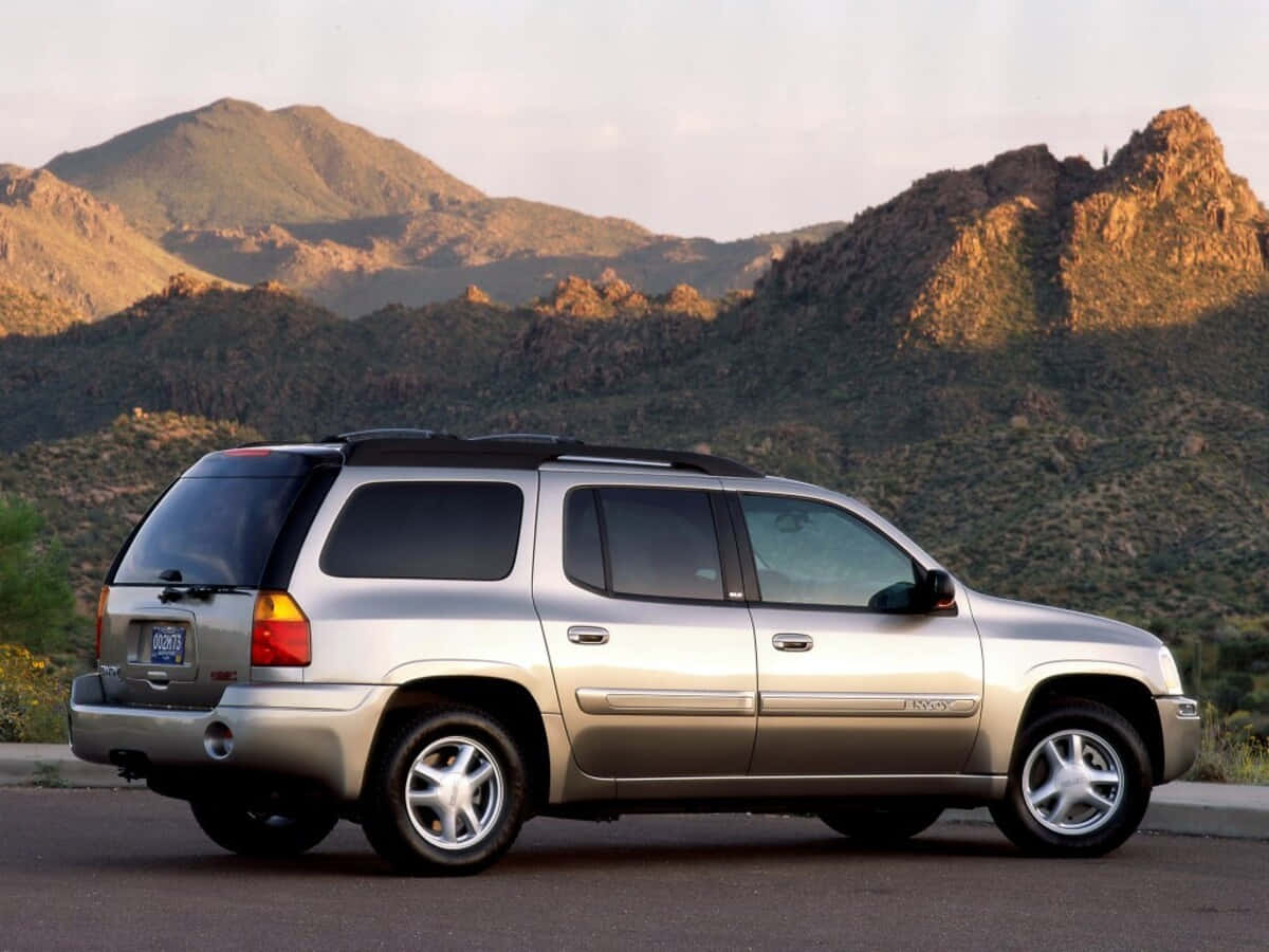 Gmc Envoy 桌布 壁紙 — 一輛銀色GMC Envoy SUV停在路邊，背景是崎嶇的山脈沐浴在金色的日落光芒中。