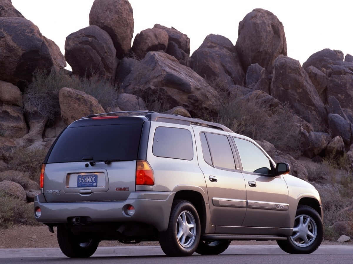 Gmc Envoy 桌布 壁紙 — 一輛銀色GMC Envoy SUV停泊在鋪砌的道路上，背景是沙漠，有巨大的粗糙岩石和乾燥的灌木。