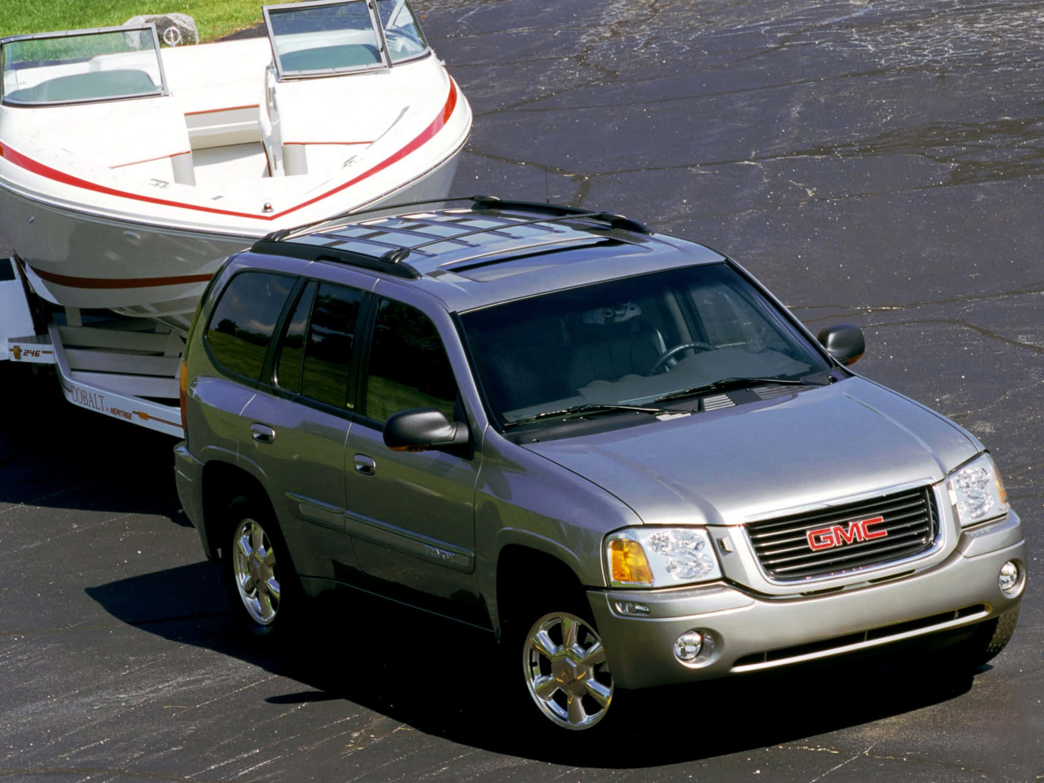 Gmc Envoy 桌布 壁紙 — 一輛銀色GMC SUV在明亮的陽光下，在裂開的瀝青路面上拖著一輛載有白色船隻的拖車。